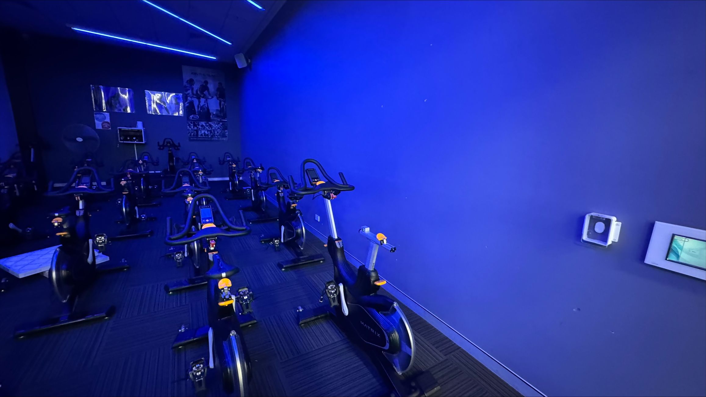 A dimly lit indoor cycling studio with a blue hue. On the left, several stationary bikes are arranged in rows, facing a dark wall with posters and a screen. The right side features a plain blue wall with a small control panel and a sensor. The ceiling has blue LED strip lights, and the floor is covered with dark carpet tiles. A dimly lit indoor cycling studio with a blue hue. On the left, several stationary bikes are arranged in rows, facing a dark wall with posters and a screen. The right side features a plain blue wall with a small control panel and a sensor. The ceiling has blue LED strip lights, and the floor is covered with dark carpet tiles.