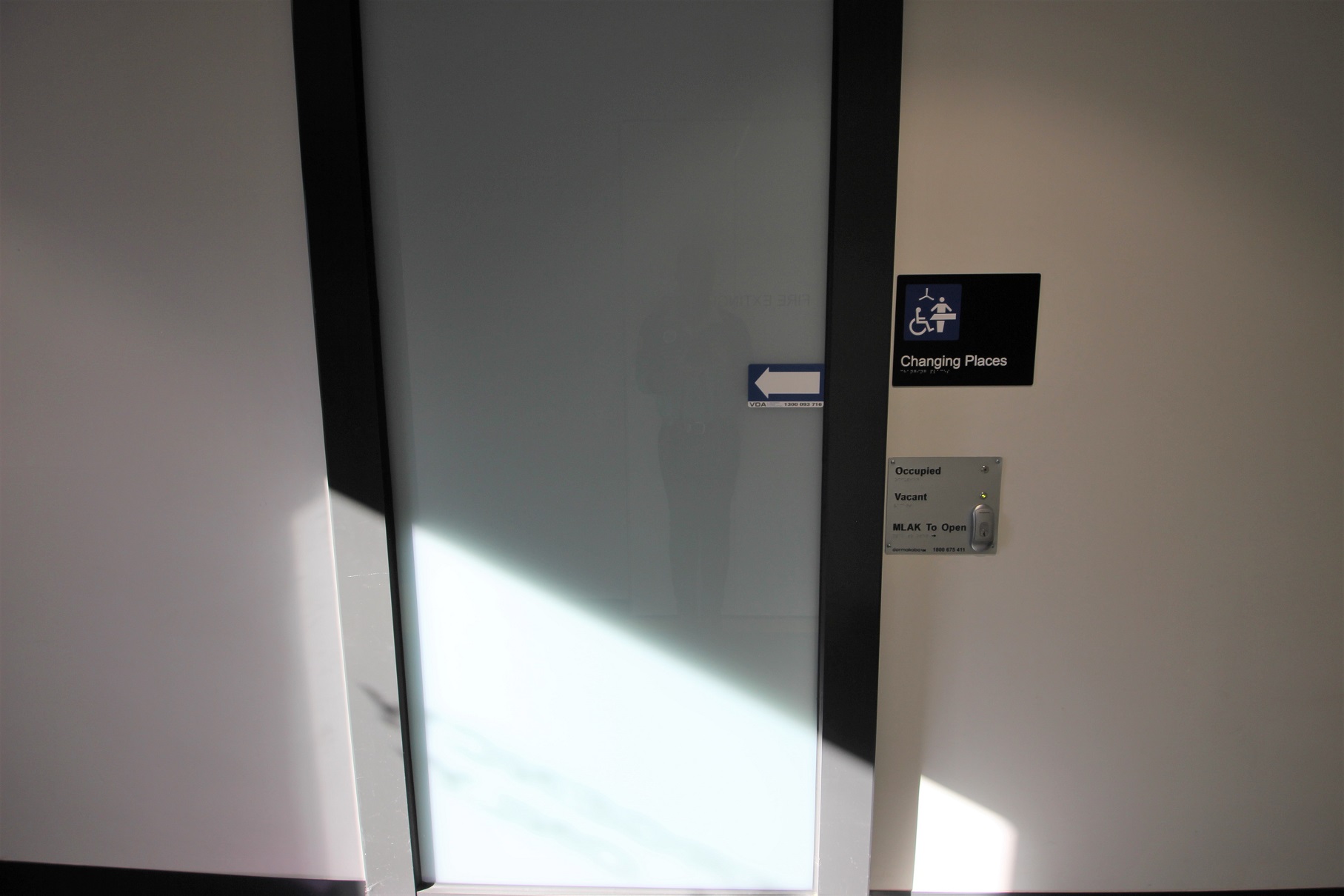 Changing places toilet at Somerville Recreation Centre. Changing places have an electronic sliding door with a push button for opening and different notification lights for occupied and vacant. A white arrow within a blue small rectangular board can be found on the grey door. There are black borders on the sides of this door.