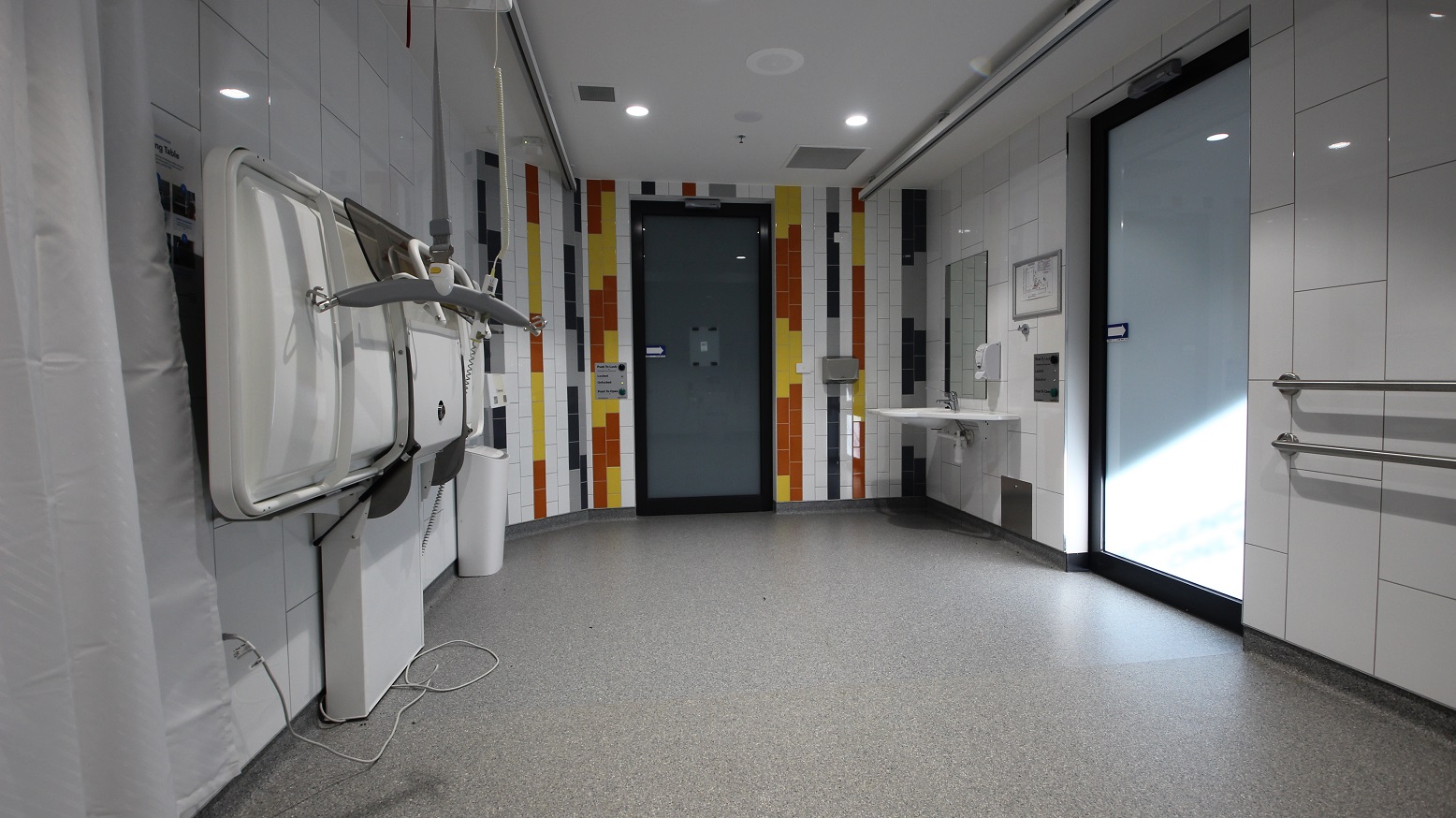 View inside the change place from the full-size white change table. There is a white bin near another electronic sliding door. The colors of the tiles on the wall near it are yellow, black, and orange. On the right side are a wash basin, an automatic liquid hand soap dispenser, a hand dryer, and a large mirror.