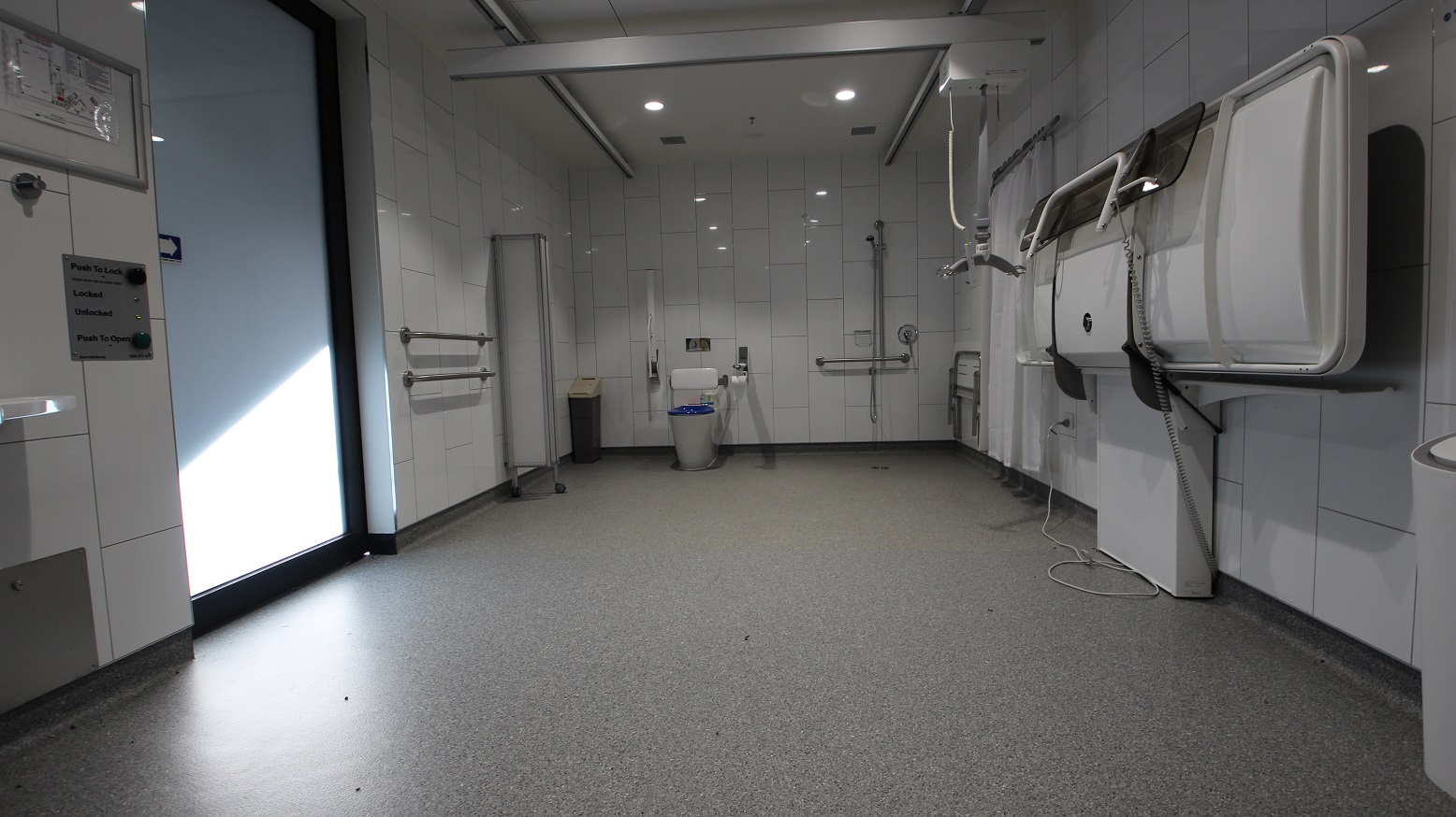Inside the change place at Somerville Recreation Centre is a shower with a white privacy curtain, grab rails, a full-size white change table, a hoist just beside the blue toilet seat with a white supporting cushion, and a white toilet roll holder. The inside looks very elegant due to its white tiled wall and granite floor.