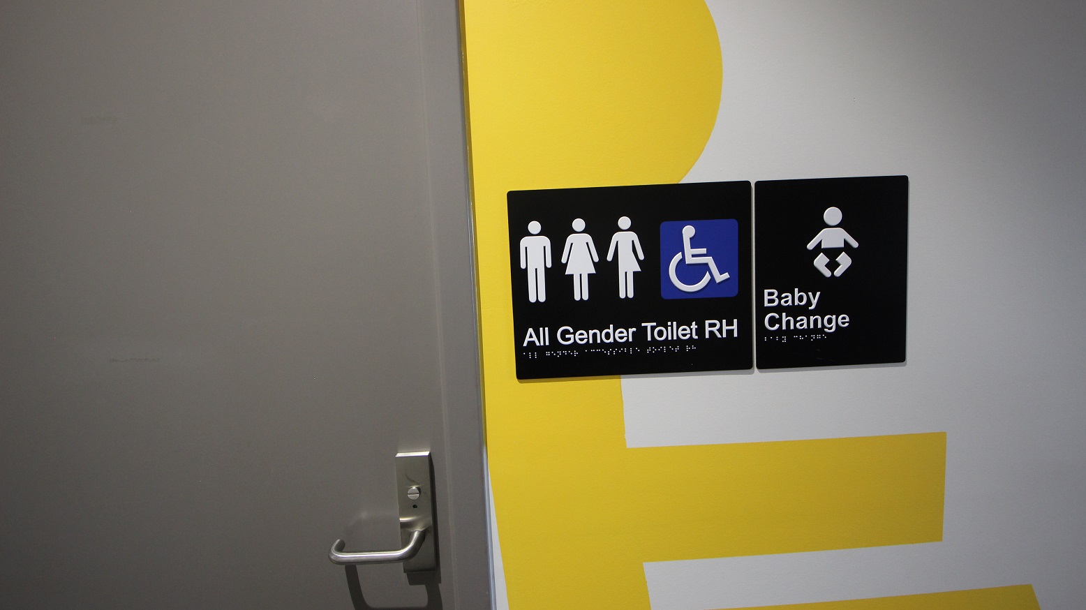 An 'All Gender Toilets' and baby change at Somerville Recreation Centre. The toilet signs have a male, female, non-binary, accessible sign, and baby icon. It has a grey door with a metal handle. There are irregular yellow patterns painted on the white wall.