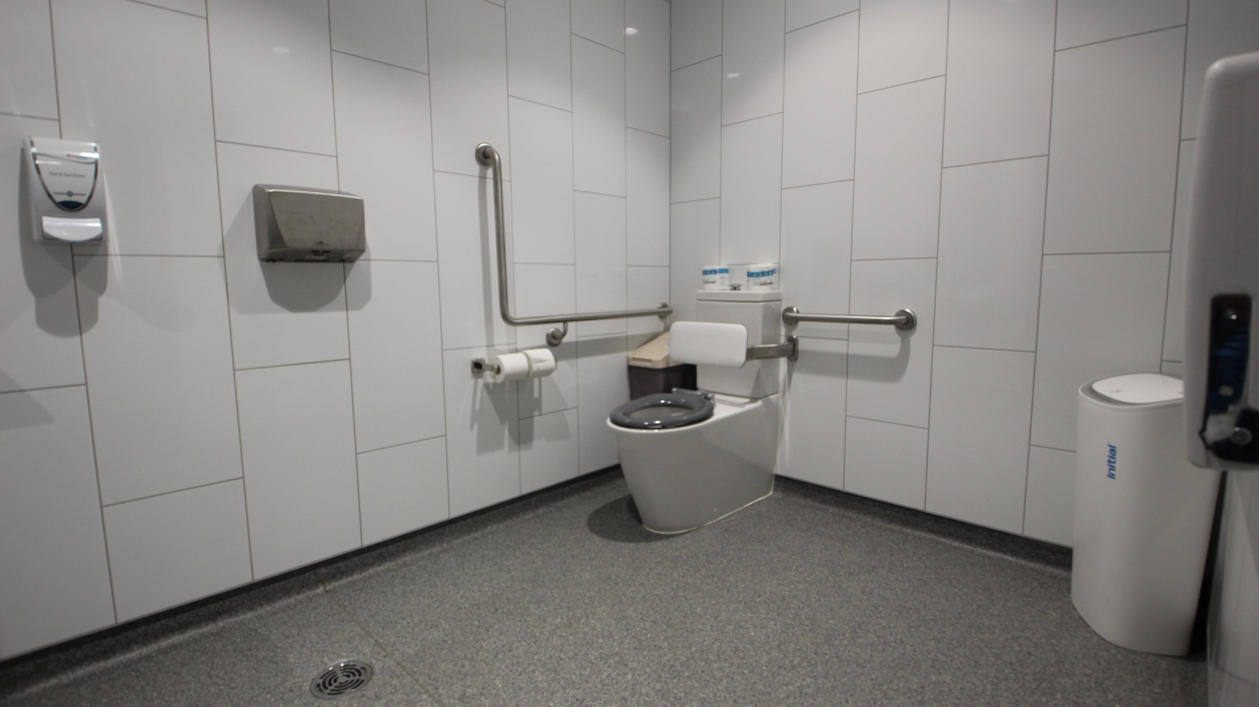Inside the “All Gender Toilets” is an accessible toilet with a white cushion for back support. On the side of the toilet seat is a brown rubbish bin with a beige lid. There are toilet papers and grab bars mounted on the white tiled wall. There is also a hand dryer and air purifier available.