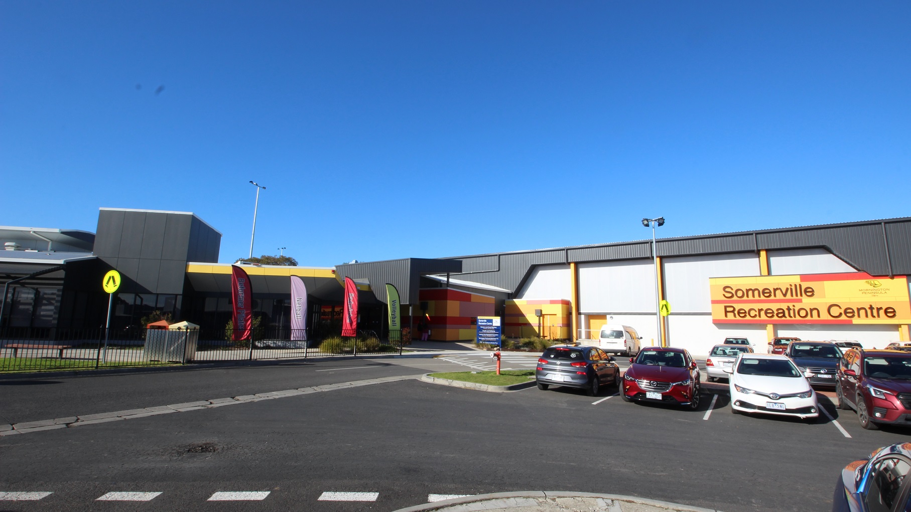Large open space car park filled with varying brands of cars. Mounted on the white front wall is the Somerville Recreation Centre board painted with their brand colors of red and yellow. Flags of different sports and colors can be seen within the hip-height length metal fences. There is also a zebra crossing from the parking to the venue.