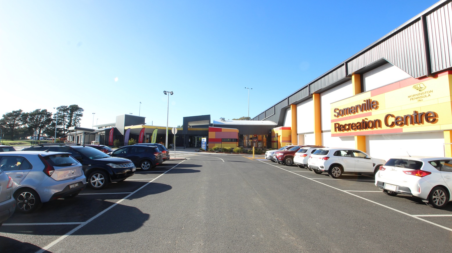 Concrete roadway in between the car spaces near the facility. At the end of this image is an orange, red, and purple painted wall. Close to the entrance, on the right, is a parking space with a blue-accessible parking sign, yellow lanes, and yellow bollards. The facility is surrounded by a beautiful green area with well-groomed landscaping and tall trees providing shade.