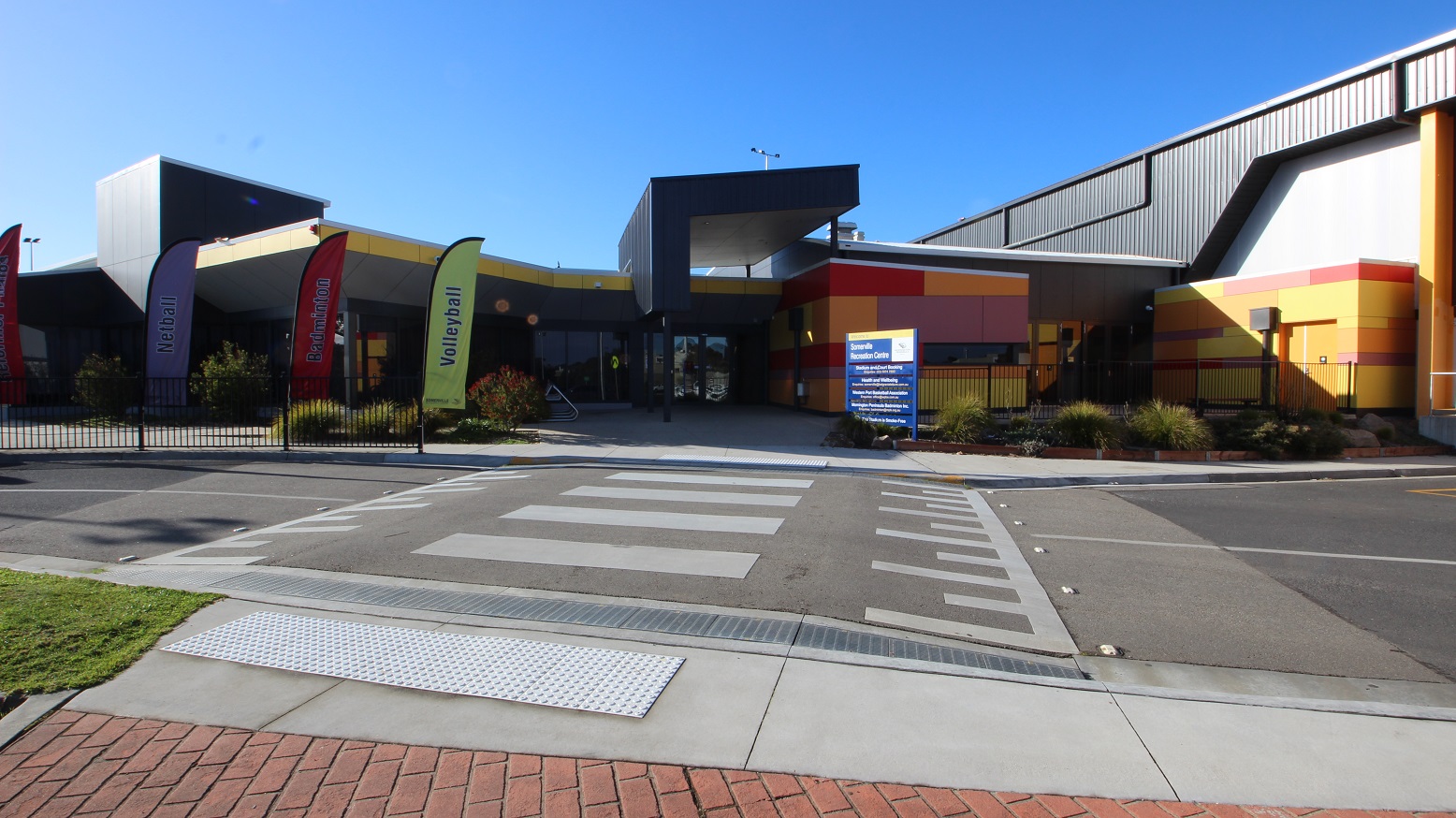 Before the zebra crossing leading to the entrance of Somerville Recreation Centre is a red brick pathway connected to a small smooth concrete section with circular metal tactile tiles. There are garden boxes filled with bushes and terrestrial plants in the concrete space right outside the building.