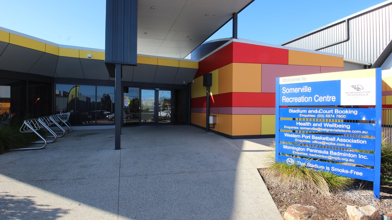 The concrete floor of the open area before the glass entrance doors has a rough-looking surface. A blue signboard with different amenities and its corresponding email for inquiries can be seen on the right side. To the left is a metal bike rack and shopping carts.