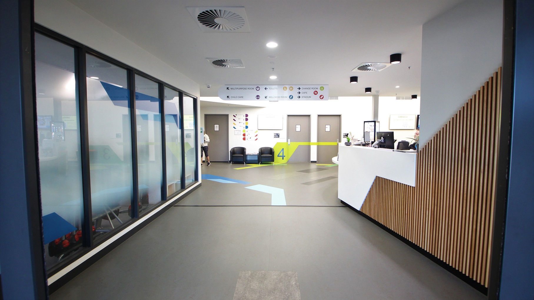 Entry to Somerville is through automatic glass, sliding doors with black metal frames. The space before the entrance door has granite flooring. On the right wall is an orange, red, and purple painted wall. The ceiling is painted in white. Reflected through the glass doors are the parking space, bike rack, flags, and pedestrian crossing.