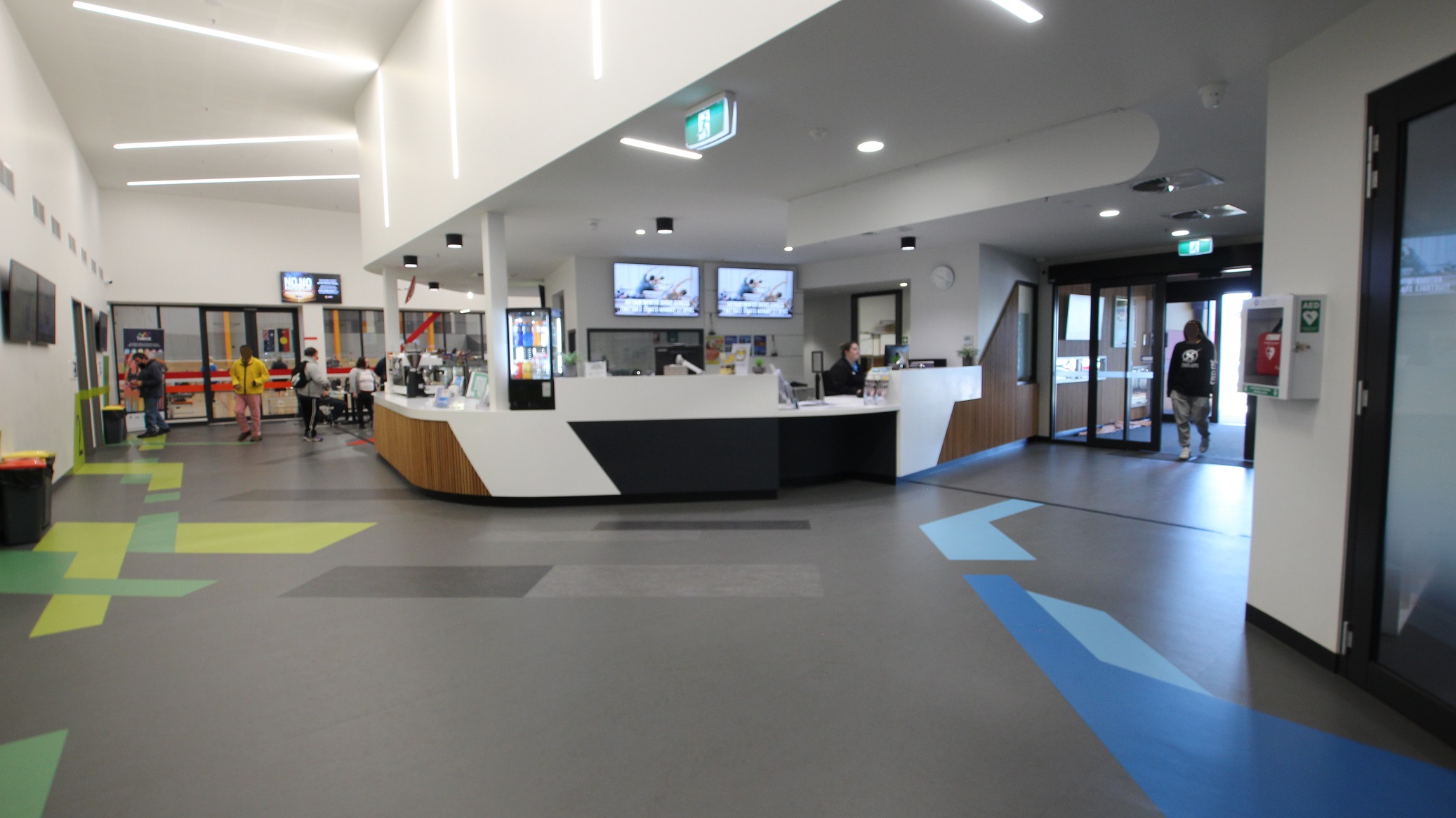 Further to the right of the reception desk is the Swell Café which has cabinets full of food and sweets as well as a coffee machine and a range of drinks. The floor has yellow-green, green, and blue and sky blue paints of irregular shapes and patterns. A number of people can be seen taking an order from the café.