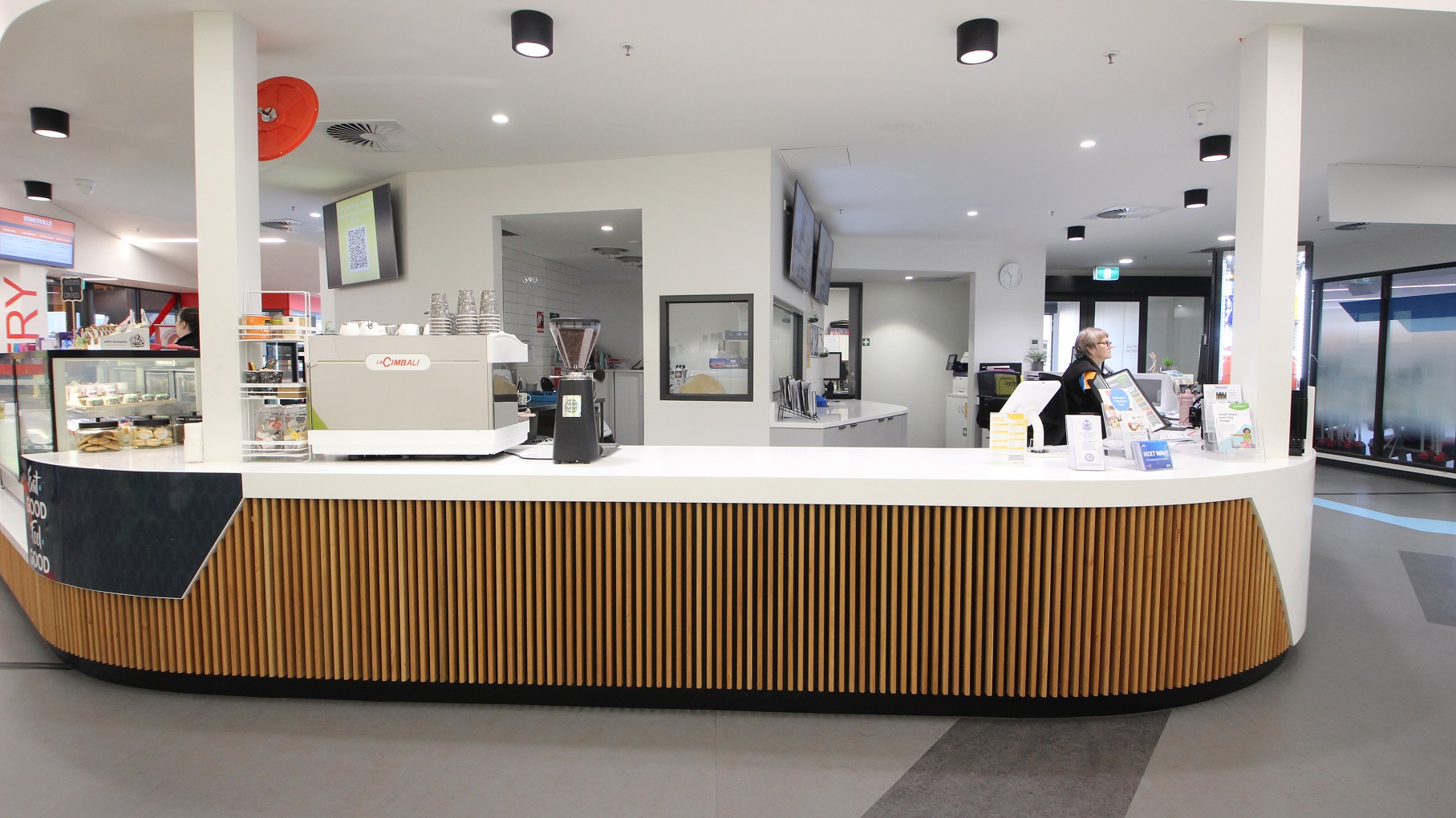 The countertop at the Swell Café has a brown wooden stick design similar to the reception desk at Somerville Recreation Centre. The staff can be identified by their black uniforms. On the upper wall are television sets. There is a separate room with a kitchen island. On the ceiling are black cylinder fluorescent lights.