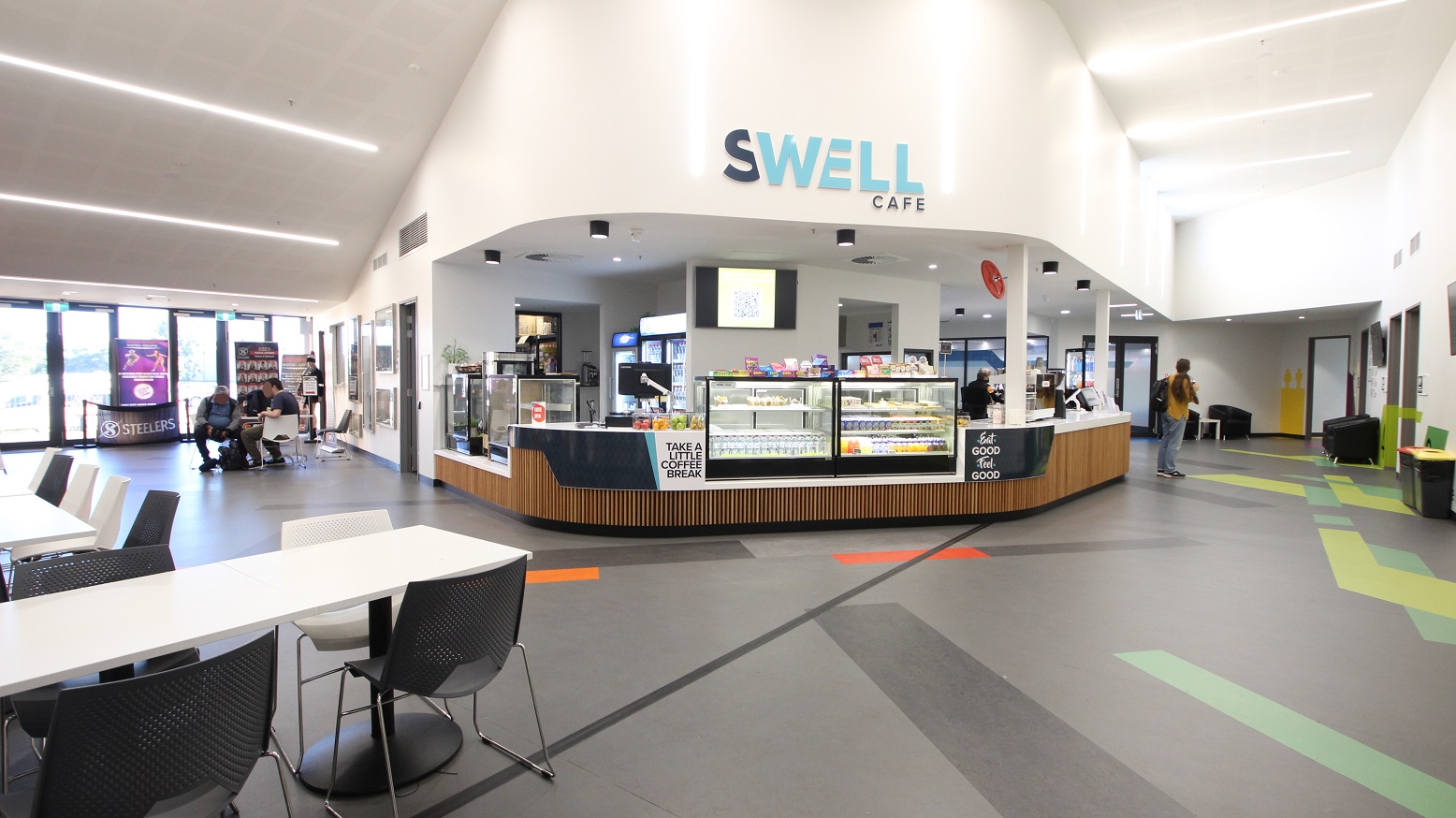 Front look of the Swell Café with its glass display cabinet full of food and sweets. On the upper wall is a Swell Café sign. Opposite the café is a seating area consisting of a white rectangular table with black chairs and steel legs. The floor has yellow-green, green, and blue and sky blue paints of irregular shapes and patterns.