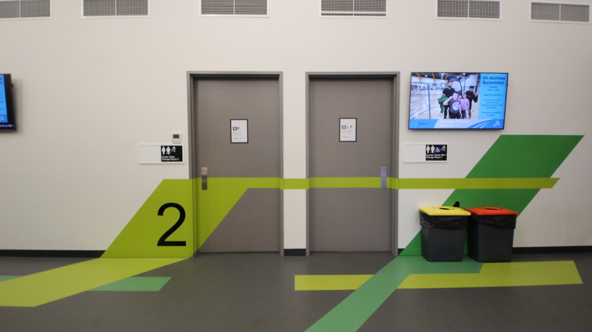 Accessible toilet and change rooms 1 and 2 available on this floor of Somerville Recreation Centre. These two floors have different hues of green paint on some parts of its door and walls. On the right are rubbish bins with orange and yellow lids. A large TV is mounted on the white wall on the right. Several exhaust hoods can be seen on the upper wall.