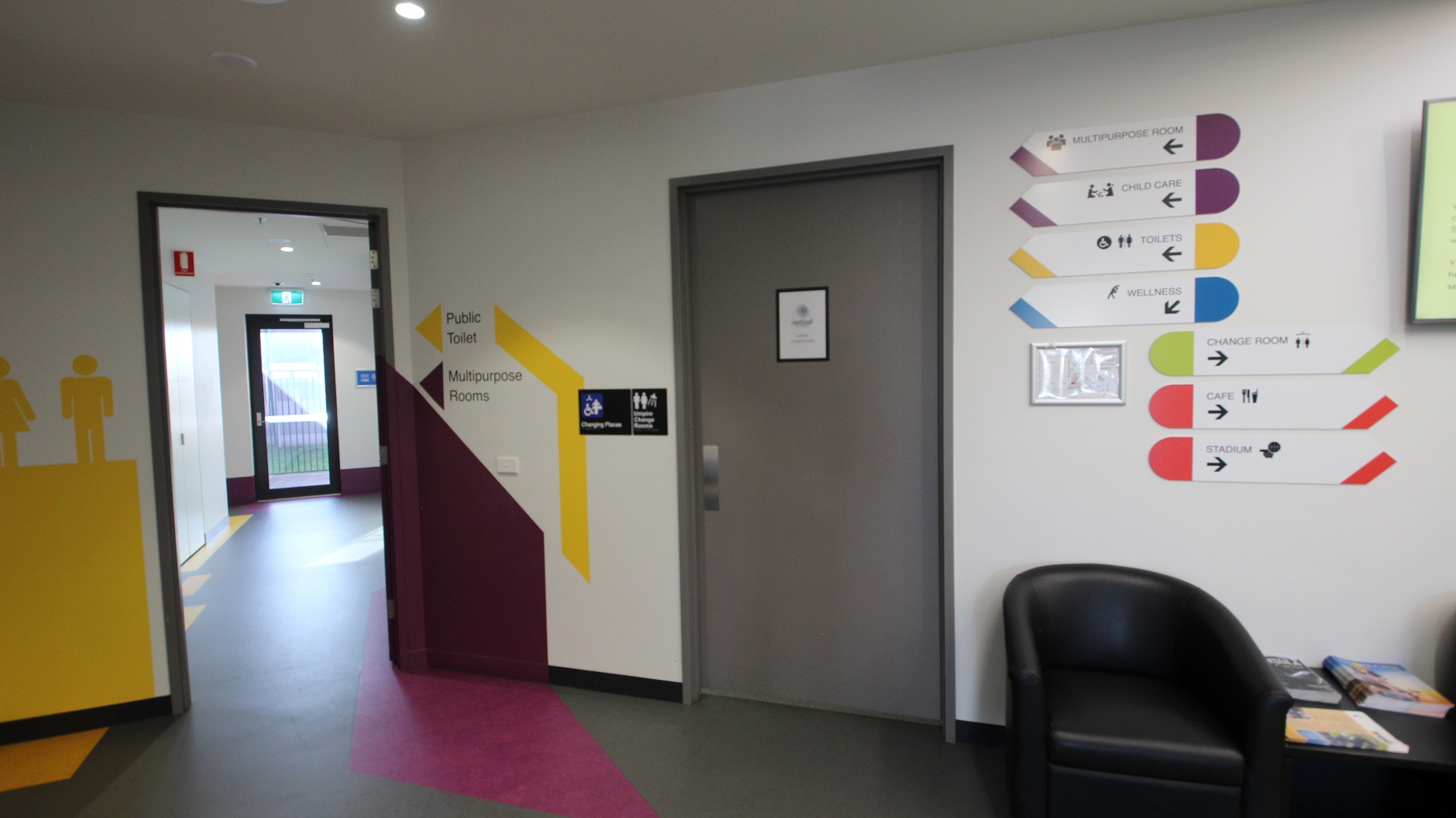 Corridor leading to the public toilets and multipurpose rooms. An accessible changing place with a grey door can also be found on this floor. On the right is a black sofa chair and on the white wall are small, white sign boards for the different amenities inside the centre.