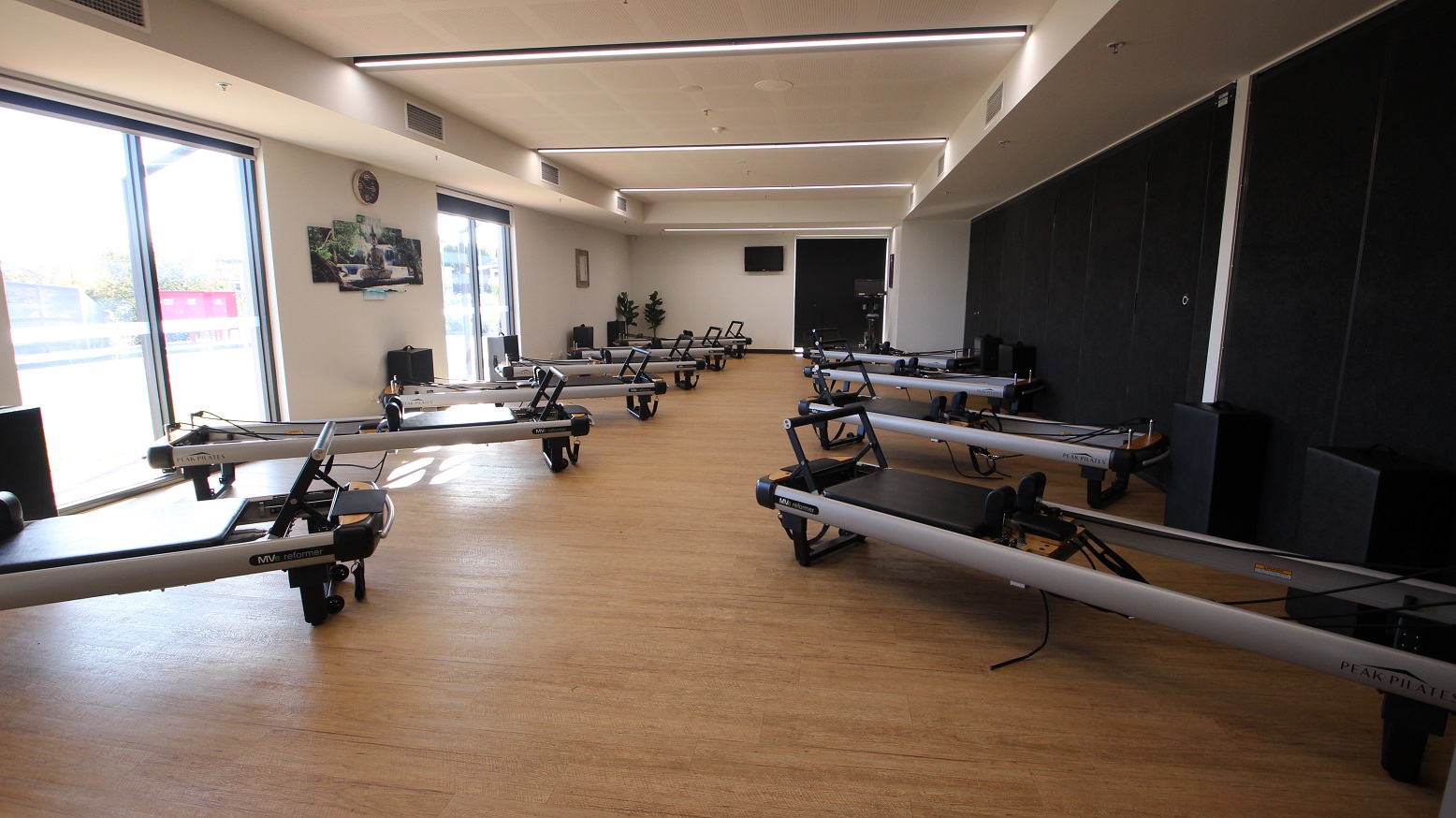 Inside of the Reformer Pilates room. It has a brown timber floor with two rows of Reformer Pilates Machines. The room has a combination of natural light filtering through the windows as well as lighting from the ceiling. Exhaust hoods are found on the upper part of the wall. There are black speakers and potted plants scattered inside the room. On the left is a black wall.