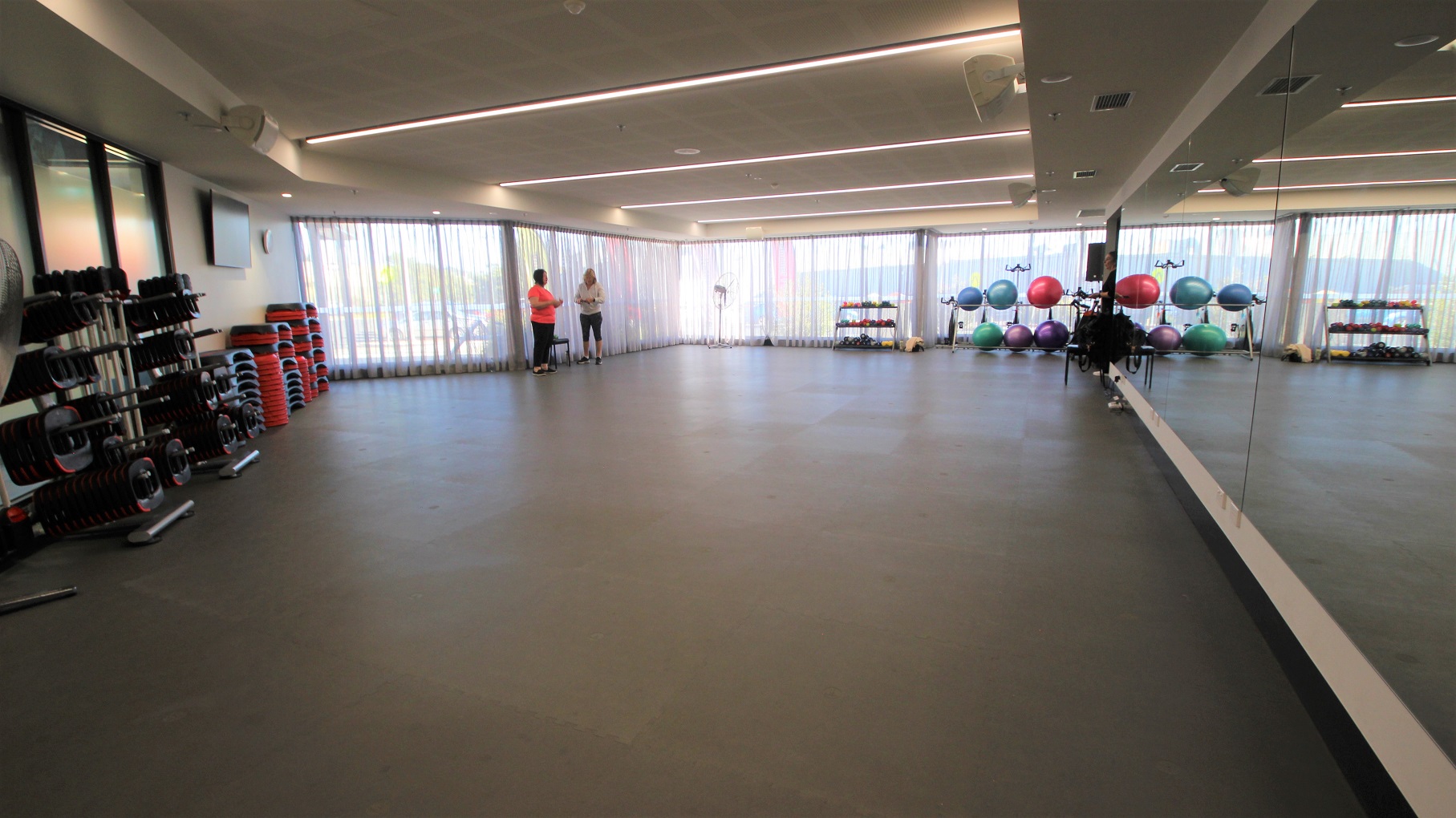 Room for Group Fitness Classes at Somerville Recreation Centre. One of its features is a floor-to-ceiling mirror in front. The glass walls are covered by white thin curtains. Found also in this room a range of weights, exercise balls, and other gym equipment. The floor is covered with a grey mat.
