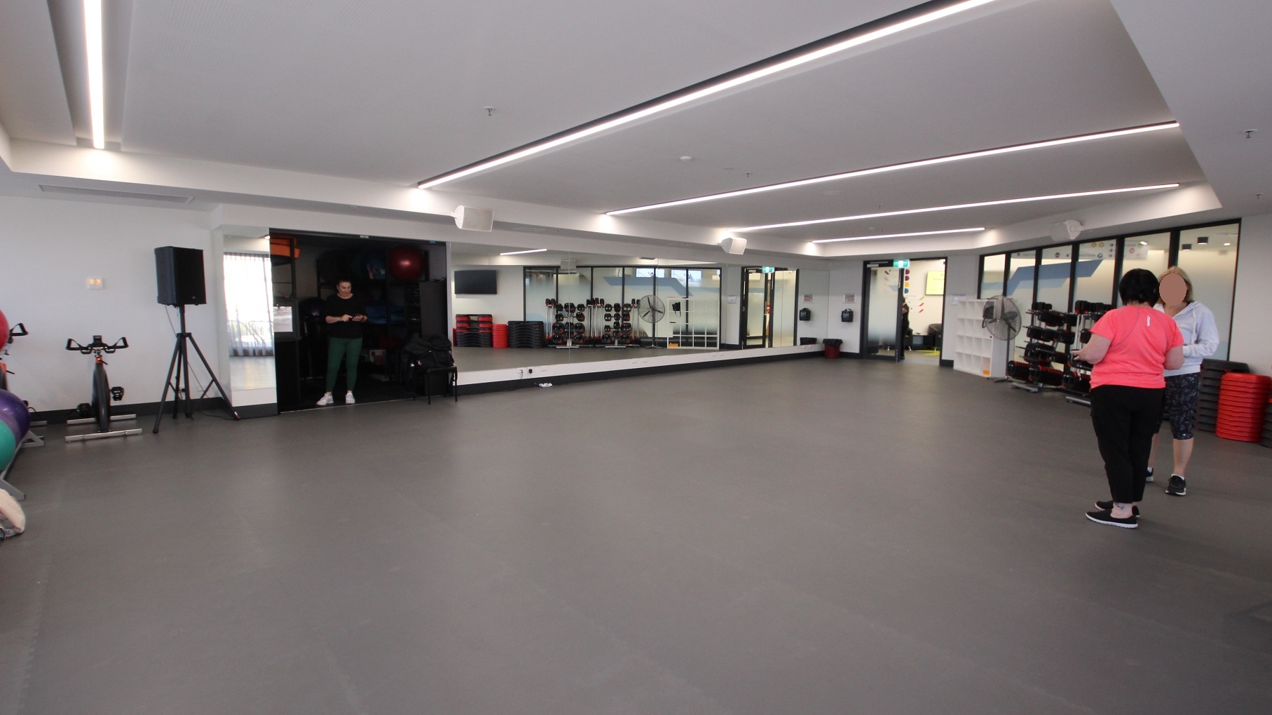View opposite the entrance door at the Group Fitness Classes room at Somerville Recreation Centre. There is a floor-to-ceiling mirror in front and next to it is a dark room and a white cabinet for bags near the glass doors. Near the gym balls are a bicycle and a speaker with a stand.