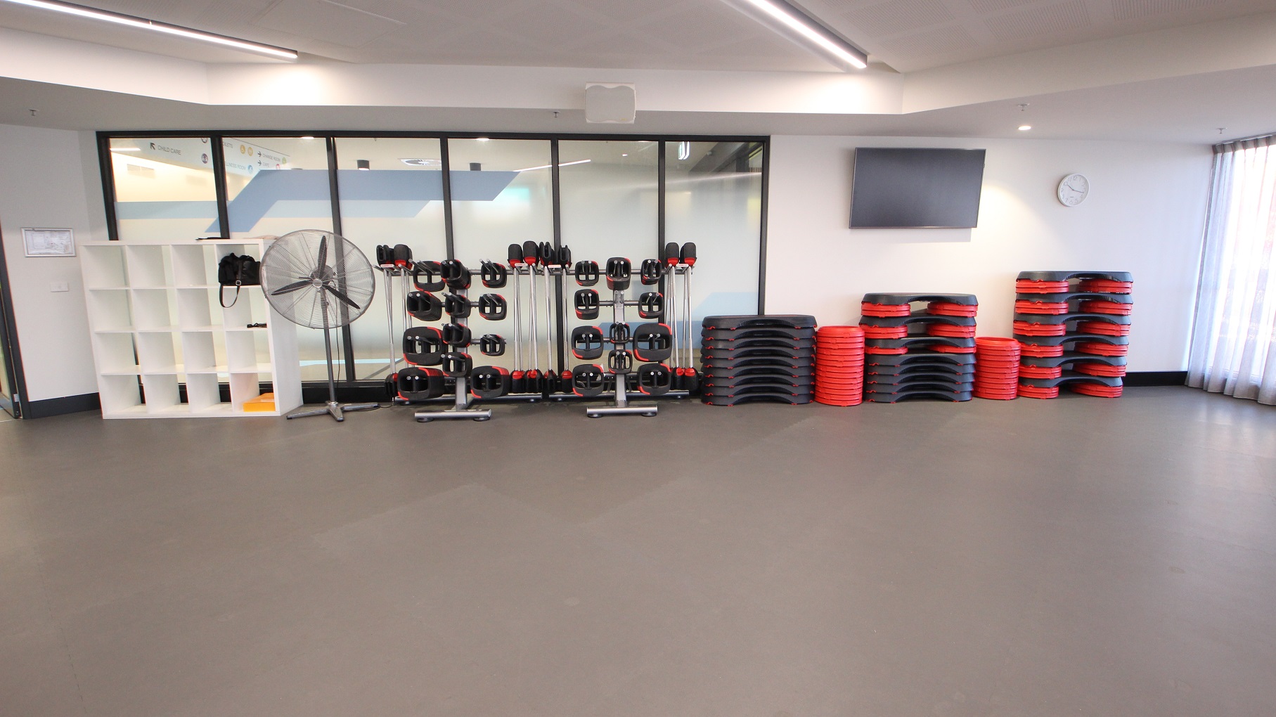 A closer look at the white cabinet with bags near the glass doors, a large stand fan, smart bars, and a red & black step riser set in the Group Fitness Classes room. On the right wall is a television and a white analog clock. The floor room is covered with a grey mat.