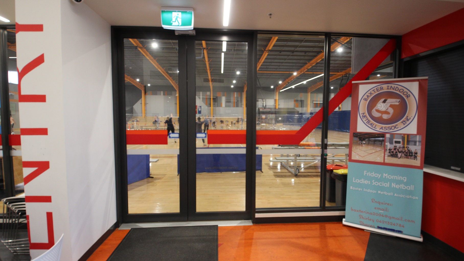 Automatic, glass sliding doors with black metal frames of the Stadium at Somerville Recreation Centre. On the right is a large marketing poster of Baxter Indoor Netball Association, Inc. Hanging on the ceiling is a neon green fire exit sign. Seen through the glass doors is the large, polished light pine timber floor of the stadium.