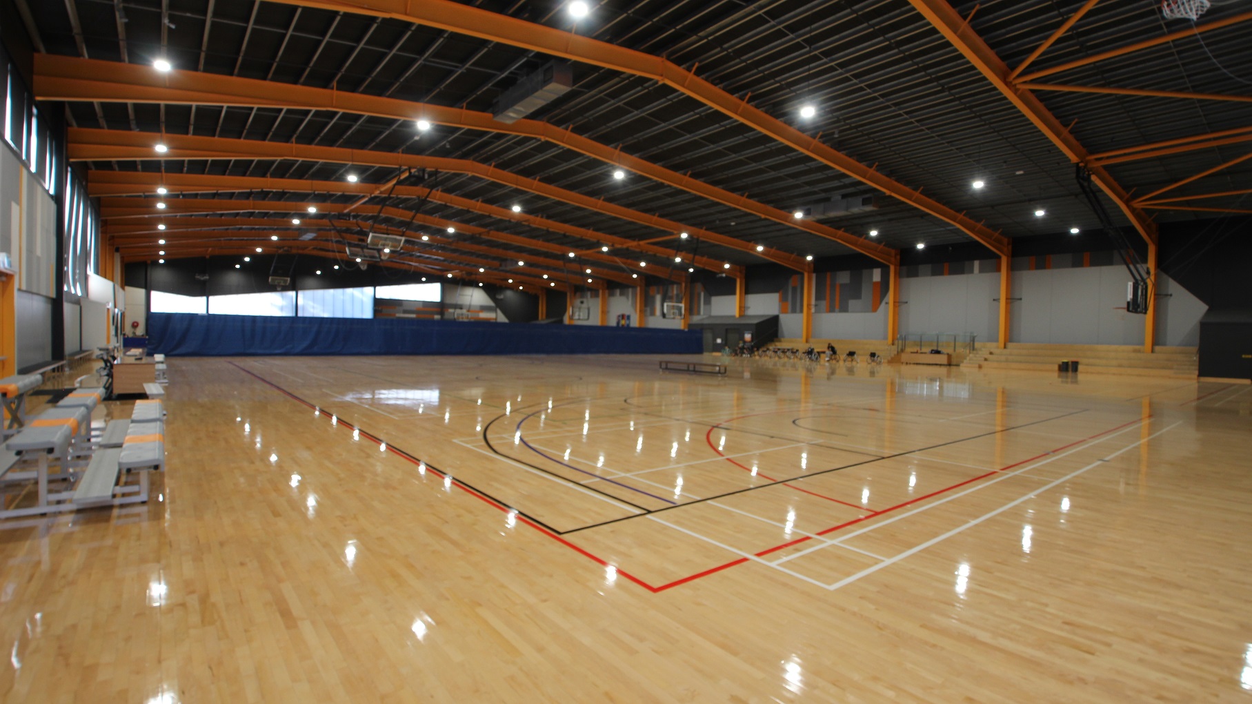 The lights coming from the fluorescents on the ceiling are seen to be reflected on the polished pine timber floor. The scorer's table is located in between the grey benches with orange lanes on the left side of the court, while on the opposite is a 4-tier seating.  The stadium has a grey ceiling and bright yellow metal archways.