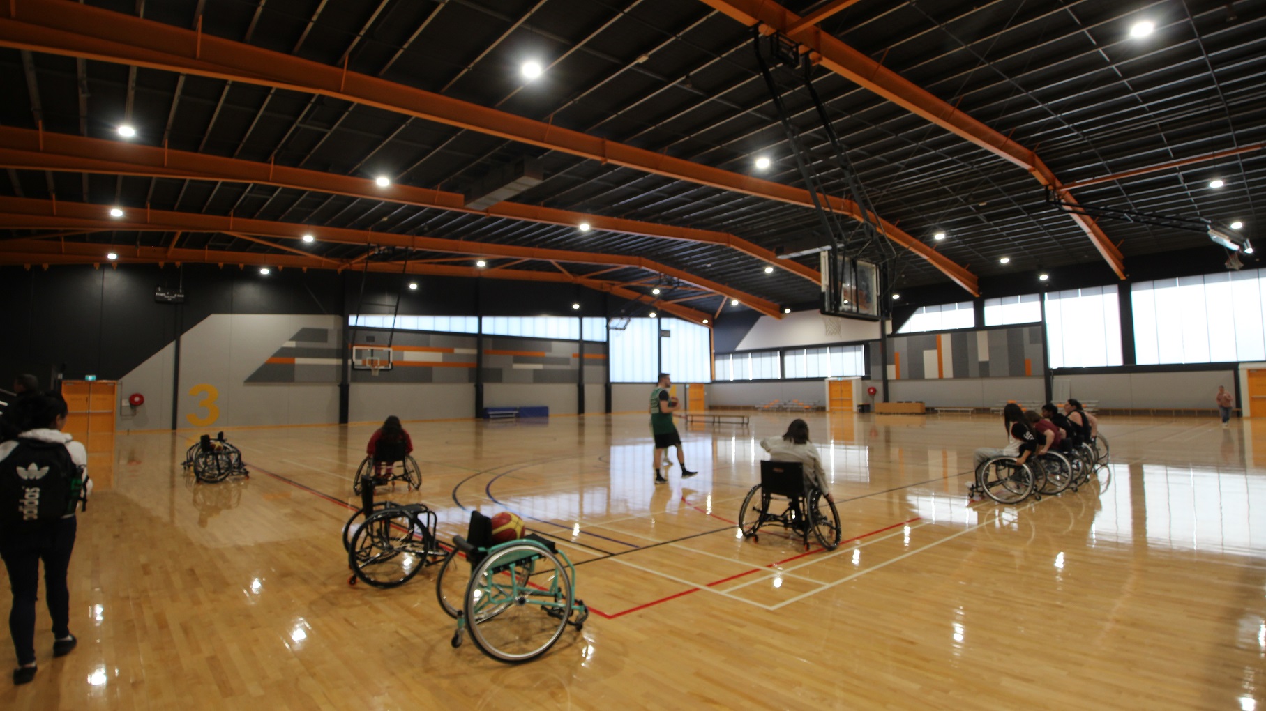 Persons with disabilities moving around the polished pine timber flooring using wheelchairs are waiting for the instruction of their basketball coach holding a ball. The grey wall at the back has a number 3 painted in orange color. Natural light can pass through the clerestory windows creating a beautiful effect on the floor.