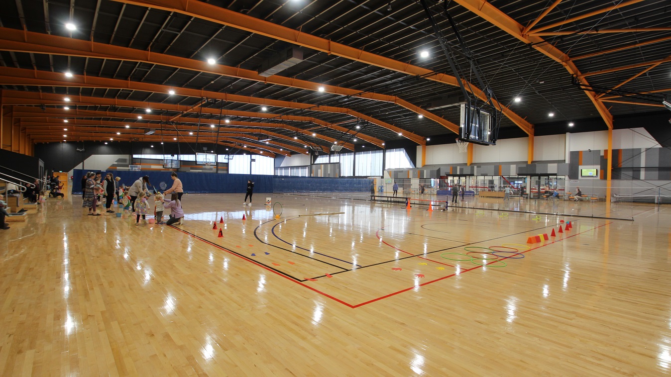 This section of the stadium separated by a plastic wall is occupied by kids and their parents playing a game using red cones, hula hoops, and cut papers. Other parents can be seen in the gymnasium seating area. On the opposite side is the badminton court.