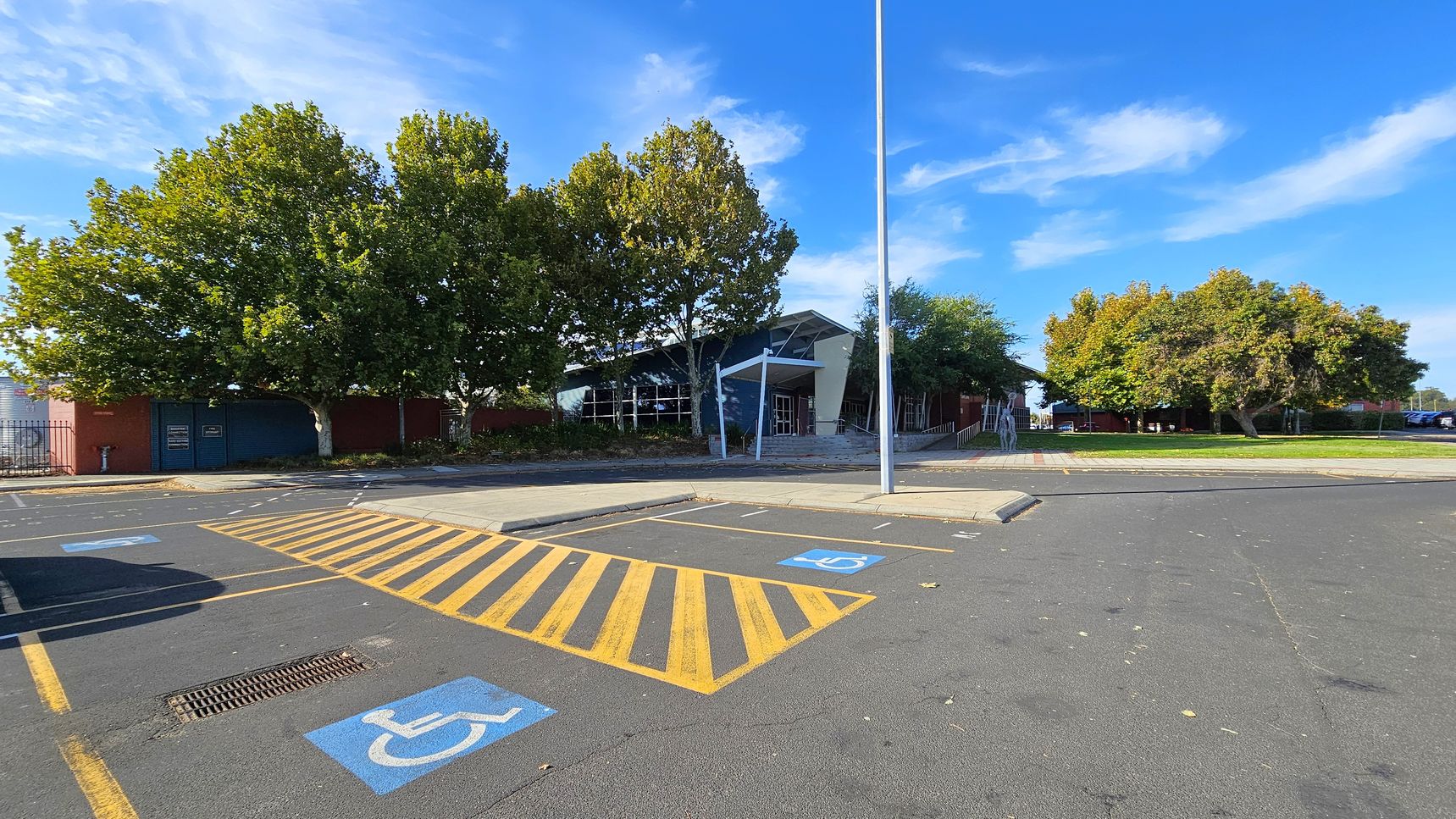 A parking area with marked disabled parking spaces in the foreground, featuring blue symbols and yellow striped lines. In the middle, a modern building with a slanted roof and large windows is partially obscured by trees. The background has more trees and a grassy area. A tall streetlight stands near the centre. The pavement is dark grey, and the sky is clear with some clouds. A parking area with marked disabled parking spaces in the foreground, featuring blue symbols and yellow striped lines. In the middle, a modern building with a slanted roof and large windows is partially obscured by trees. The background has more trees and a grassy area. A tall streetlight stands near the centre. The pavement is dark grey, and the sky is clear with some clouds.