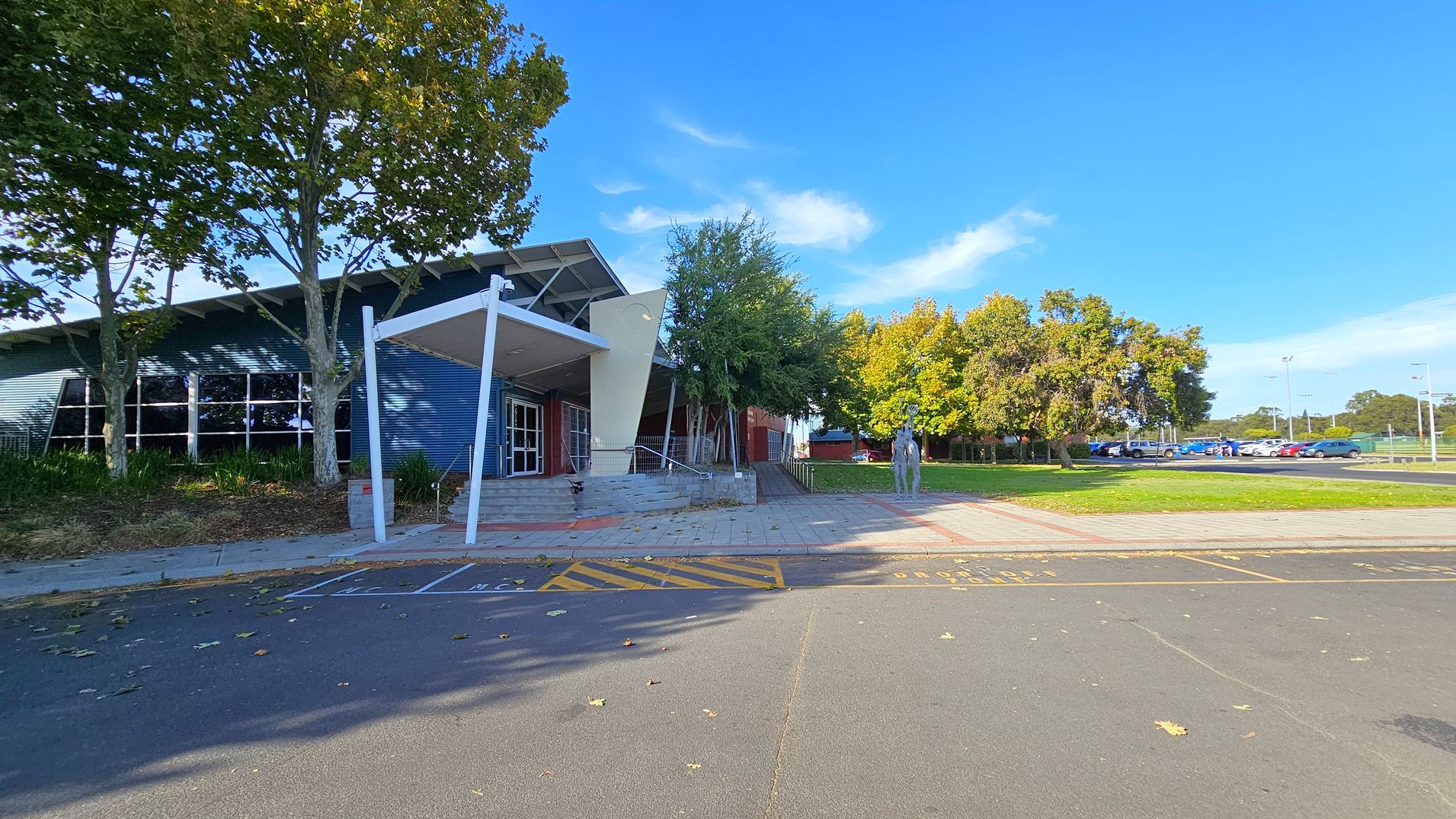 A modern building with a slanted roof and large windows is on the left, partially shaded by tall trees. The entrance features a white canopy and steps leading up to it. On the right, a paved area with a sculpture of a person and more trees in the background. A car park with several vehicles is visible further right. The ground is a mix of asphalt and paving, with some fallen leaves scattered around. A modern building with a slanted roof and large windows is on the left, partially shaded by tall trees. The entrance features a white canopy and steps leading up to it. On the right, a paved area with a sculpture of a person and more trees in the background. A car park with several vehicles is visible further right. The ground is a mix of asphalt and paving, with some fallen leaves scattered around.