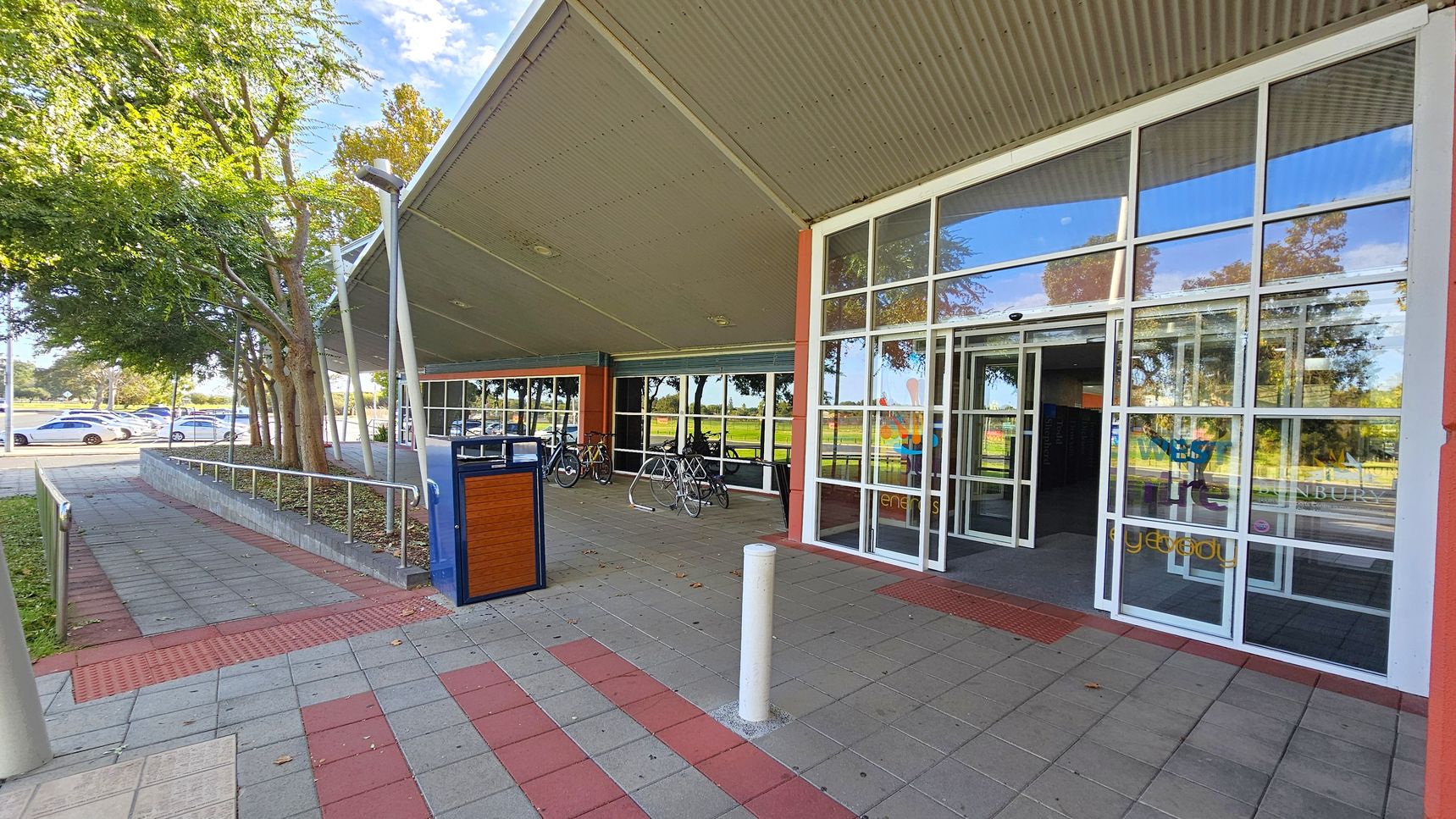 A building entrance with large glass doors and windows on the right. The foreground has a tiled pavement with red and grey patterns. A white bollard and a blue and wooden bin are near the entrance. On the left, there are trees and a row of parked cars. Bicycles are parked under a covered area with a slanted roof. The background shows more trees and a grassy area. A building entrance with large glass doors and windows on the right. The foreground has a tiled pavement with red and grey patterns. A white bollard and a blue and wooden bin are near the entrance. On the left, there are trees and a row of parked cars. Bicycles are parked under a covered area with a slanted roof. The background shows more trees and a grassy area.