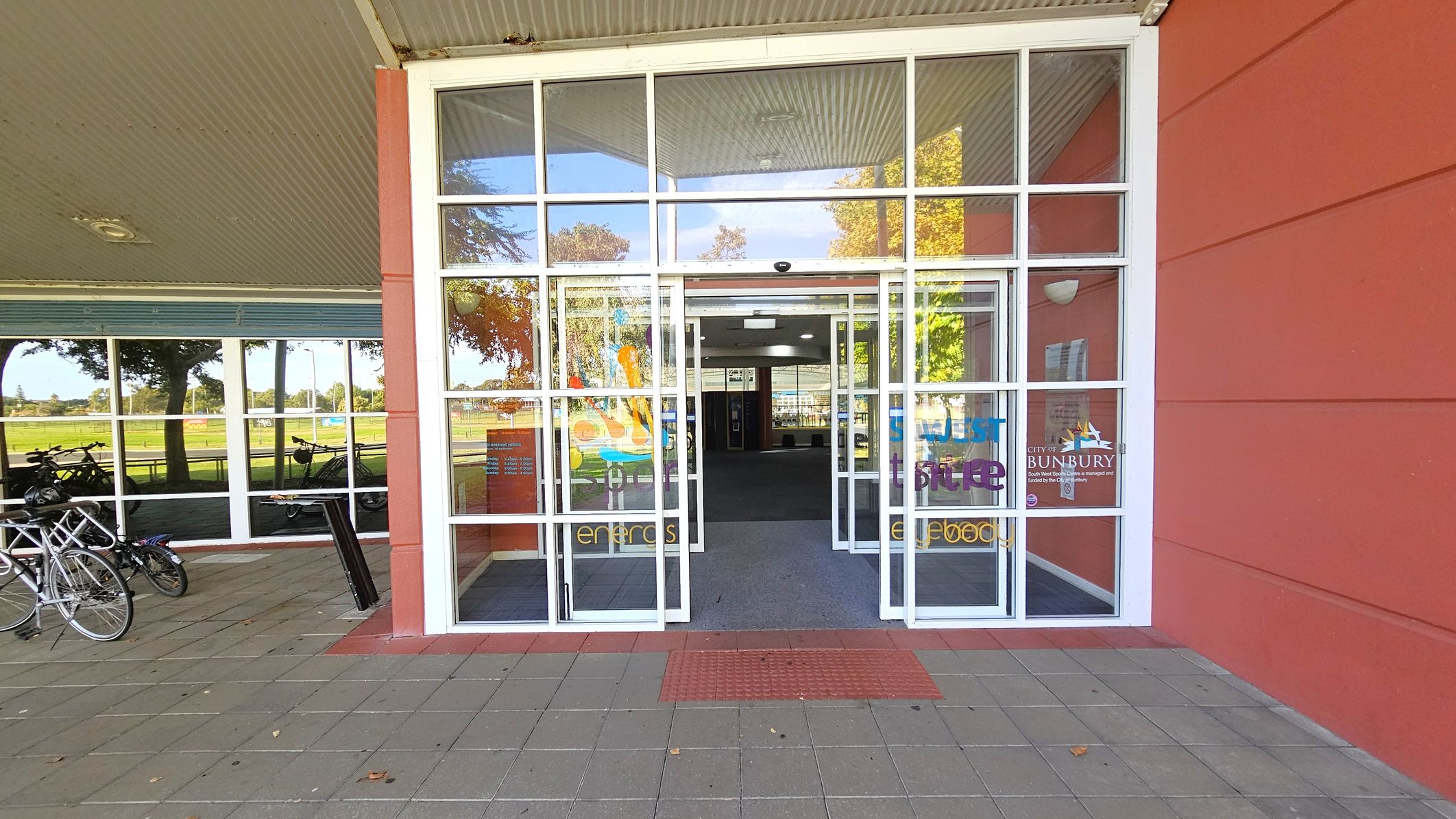 A glass entrance with double doors is centred, featuring colourful text and logos. The building has a red exterior wall on the right. On the left, bicycles are parked against a window reflecting trees and a grassy area. The floor is paved with grey and red tiles, and a red tactile mat is in front of the entrance. The ceiling is corrugated metal with a light fixture. A glass entrance with double doors is centred, featuring colourful text and logos. The building has a red exterior wall on the right. On the left, bicycles are parked against a window reflecting trees and a grassy area. The floor is paved with grey and red tiles, and a red tactile mat is in front of the entrance. The ceiling is corrugated metal with a light fixture.