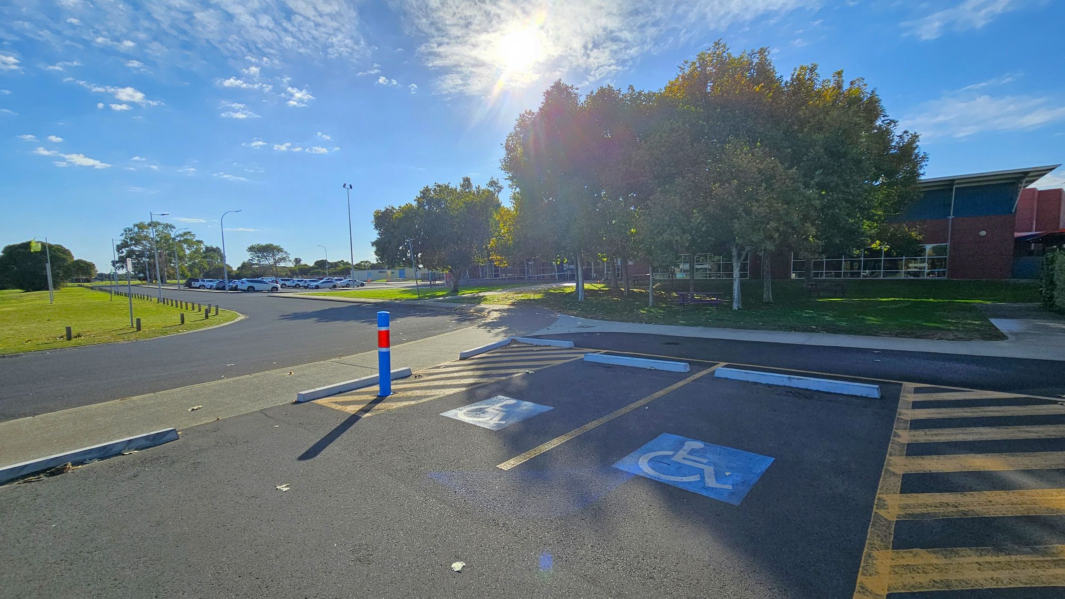 A parking area with two marked disabled parking spaces in the foreground. The ground is asphalt with painted symbols. A blue and red bollard is near the spaces. In the background, there are trees casting shadows and a building with large windows. To the left, a grassy area with a row of parked cars along a road. The sun is shining brightly, creating lens flare effects. A parking area with two marked disabled parking spaces in the foreground. The ground is asphalt with painted symbols. A blue and red bollard is near the spaces. In the background, there are trees casting shadows and a building with large windows. To the left, a grassy area with a row of parked cars along a road. The sun is shining brightly, creating lens flare effects.