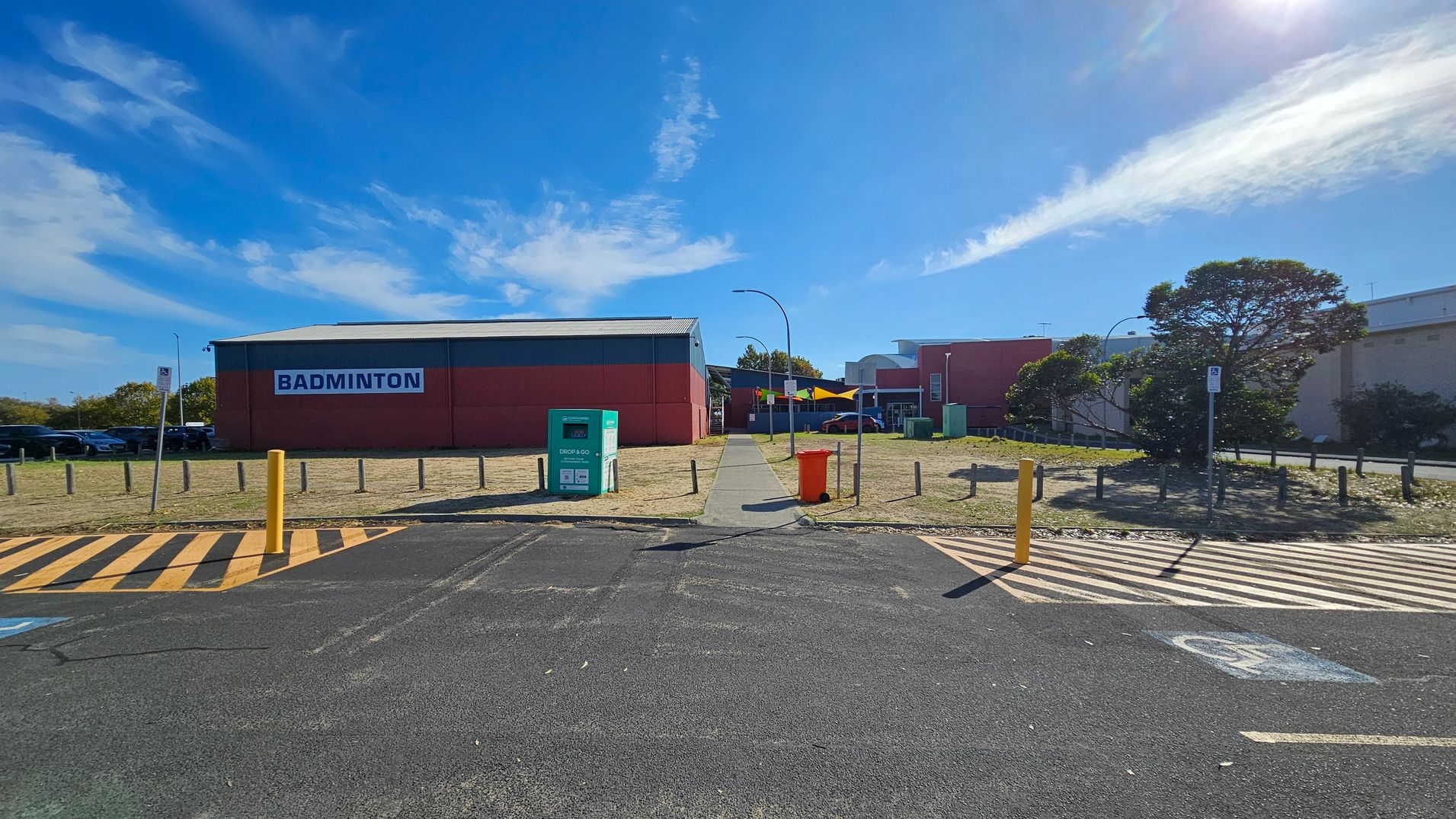 A paved pathway leads to a large red building with a sign reading "BADMINTON" on the left. In the foreground, there is a parking area with yellow and black striped markings and a yellow bollard. A green "DROP & GO" bin and an orange bin are near the path. On the right, a tree and more buildings are visible. The area is bordered by short wooden posts, and a few cars are parked on the left side. A paved pathway leads to a large red building with a sign reading "BADMINTON" on the left. In the foreground, there is a parking area with yellow and black striped markings and a yellow bollard. A green "DROP & GO" bin and an orange bin are near the path. On the right, a tree and more buildings are visible. The area is bordered by short wooden posts, and a few cars are parked on the left side.