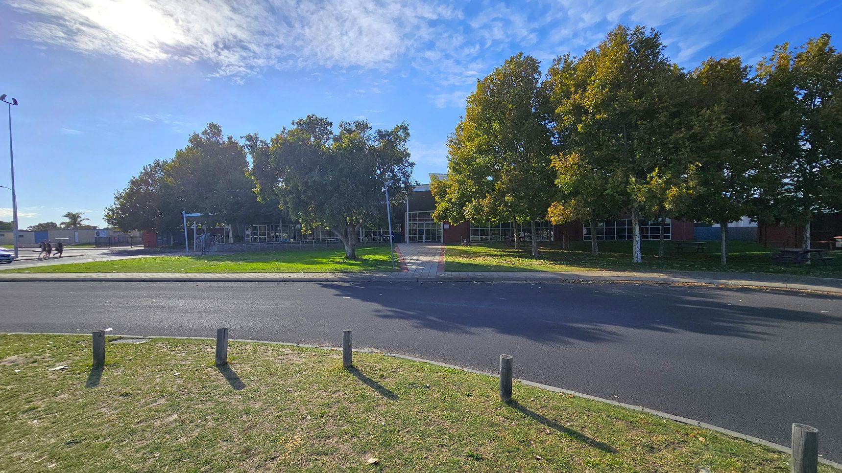 A paved road runs horizontally across the foreground with a grassy verge and wooden posts. In the middle, a path leads through a row of trees with green and yellow leaves, towards a building with large windows. On the left, a few people walk near a lamppost, and a car is parked nearby. The background features more trees and a clear sky. A paved road runs horizontally across the foreground with a grassy verge and wooden posts. In the middle, a path leads through a row of trees with green and yellow leaves, towards a building with large windows. On the left, a few people walk near a lamppost, and a car is parked nearby. The background features more trees and a clear sky.