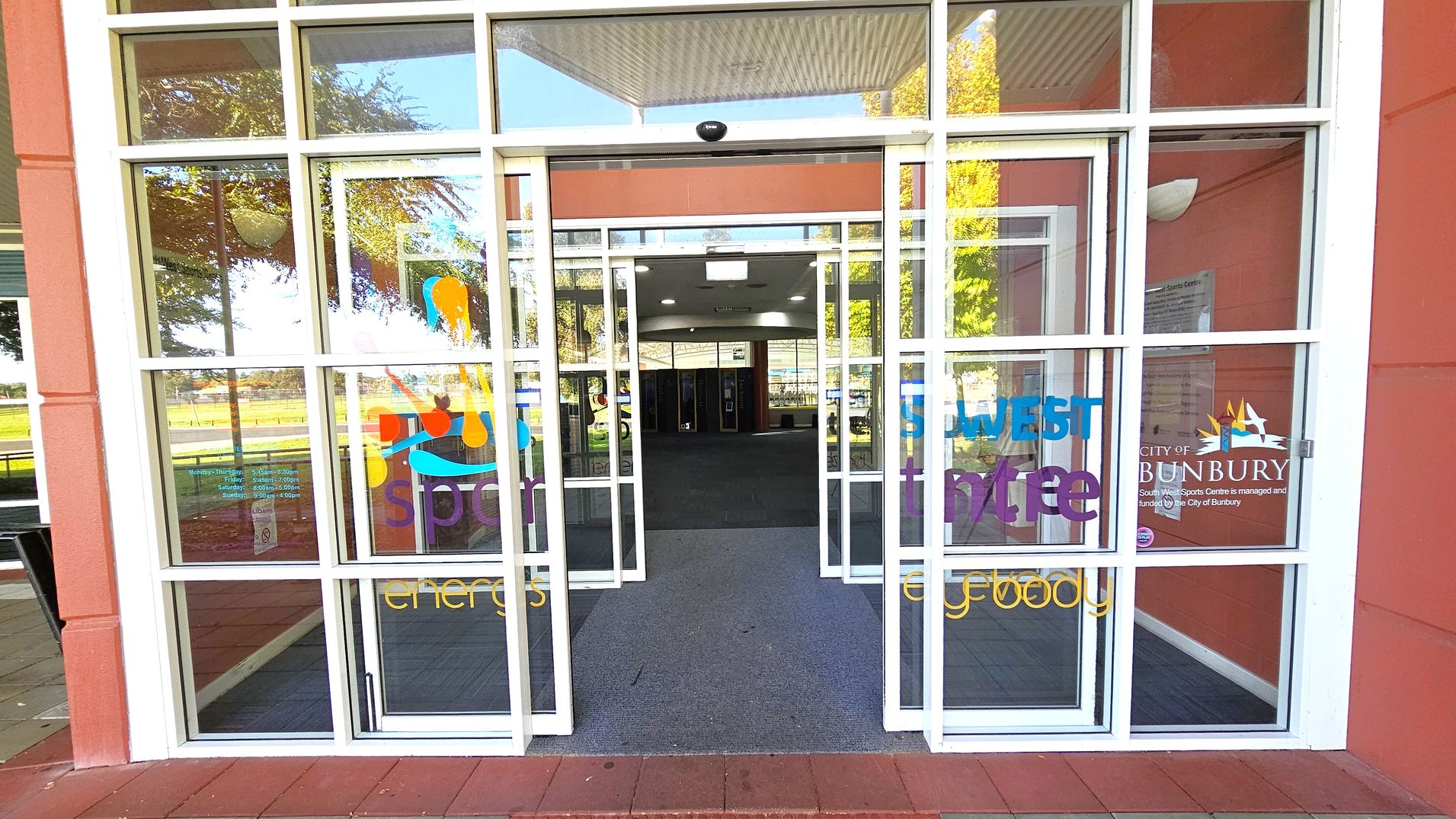 Glass entrance doors with white frames, featuring colourful text and graphics. The words "South West Sports Centre" and "City of Bunbury" are visible. Inside, a carpeted floor leads to a spacious interior with ceiling lights. Reflections of trees and outdoor seating are visible on the glass. The exterior walls are a reddish-brown colour. Glass entrance doors with white frames, featuring colourful text and graphics. The words "South West Sports Centre" and "City of Bunbury" are visible. Inside, a carpeted floor leads to a spacious interior with ceiling lights. Reflections of trees and outdoor seating are visible on the glass. The exterior walls are a reddish-brown colour.