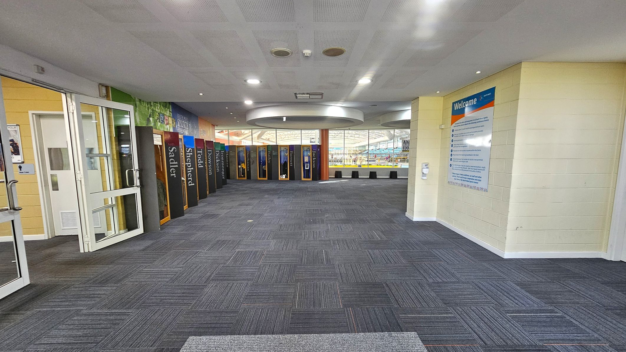 A spacious indoor area with a grey carpeted floor. On the left, there are glass doors and a sign indicating restrooms. The right side features a large welcome sign on a light-coloured wall. In the centre, a series of colourful lockers line the wall, each with a name. The ceiling is white with recessed lighting and circular fixtures. In the background, large windows reveal a glimpse of an indoor pool area. A spacious indoor area with a grey carpeted floor. On the left, there are glass doors and a sign indicating restrooms. The right side features a large welcome sign on a light-coloured wall. In the centre, a series of colourful lockers line the wall, each with a name. The ceiling is white with recessed lighting and circular fixtures. In the background, large windows reveal a glimpse of an indoor pool area.