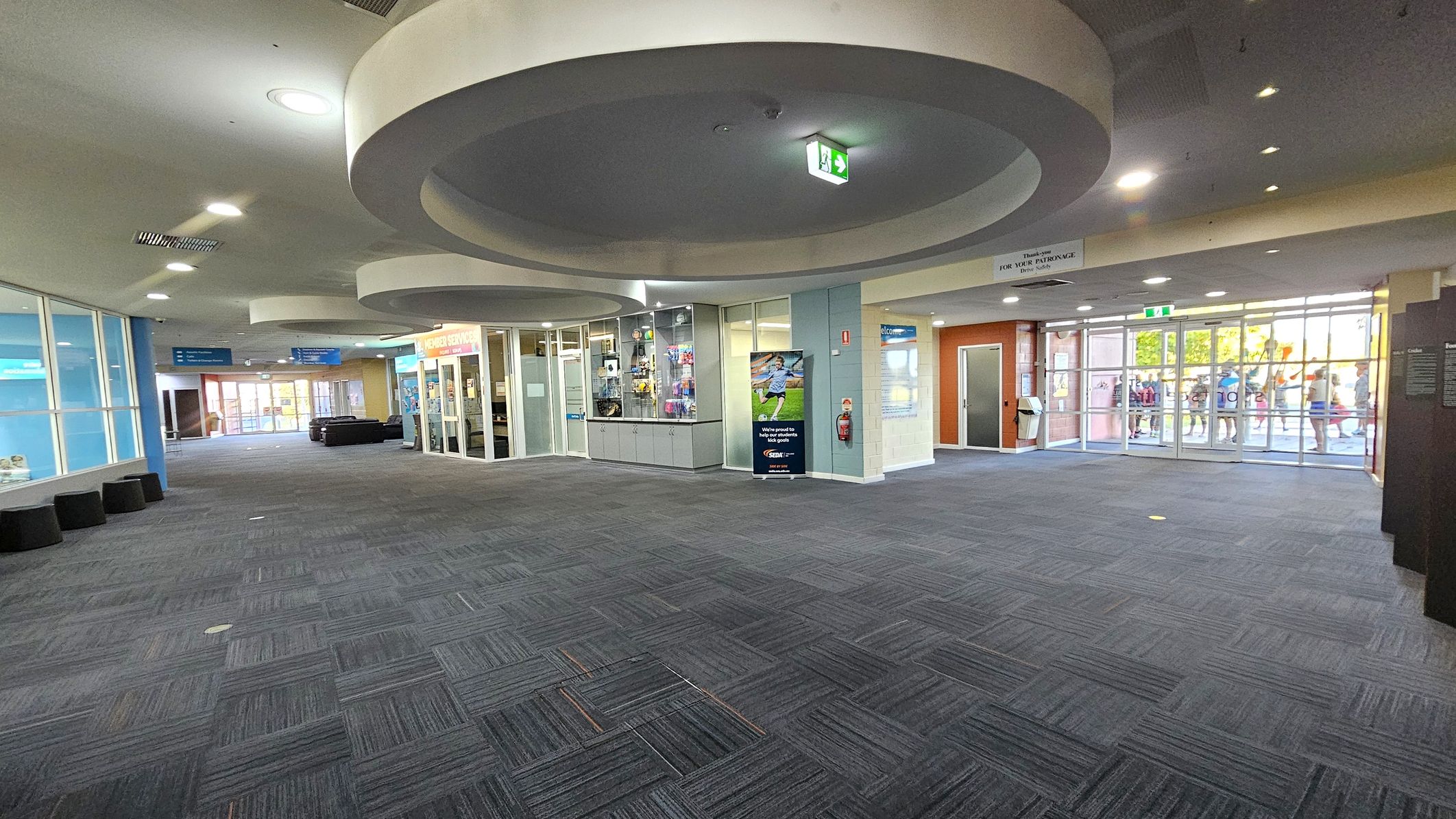 A spacious indoor area with a grey carpeted floor. On the left, there are large glass windows and a row of black cylindrical seats. In the centre, a reception area with glass walls displays various items. Circular ceiling features with recessed lighting are visible above. On the right, glass doors lead outside, where a group of people can be seen. The walls are a mix of light blue and orange tones, and there are signs and posters throughout the space. A spacious indoor area with a grey carpeted floor. On the left, there are large glass windows and a row of black cylindrical seats. In the centre, a reception area with glass walls displays various items. Circular ceiling features with recessed lighting are visible above. On the right, glass doors lead outside, where a group of people can be seen. The walls are a mix of light blue and orange tones, and there are signs and posters throughout the space.