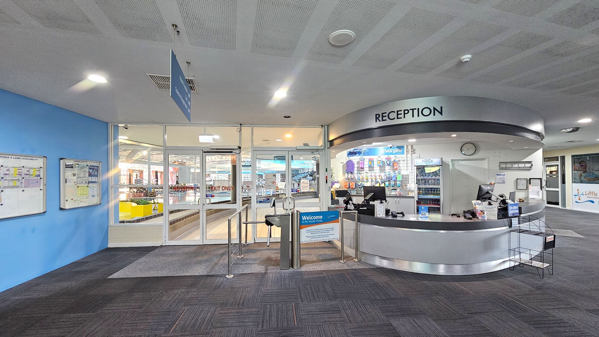 Reception area with a curved desk on the right, featuring computers, brochures, and a display of swim-related products. A sign above reads "RECEPTION". In the centre, glass doors lead to a pool area, with a turnstile and a welcome sign in front. On the left, a blue wall displays notice boards with information. The floor is carpeted in dark grey, and the ceiling is white with recessed lighting. Reception area with a curved desk on the right, featuring computers, brochures, and a display of swim-related products. A sign above reads "RECEPTION". In the centre, glass doors lead to a pool area, with a turnstile and a welcome sign in front. On the left, a blue wall displays notice boards with information. The floor is carpeted in dark grey, and the ceiling is white with recessed lighting.