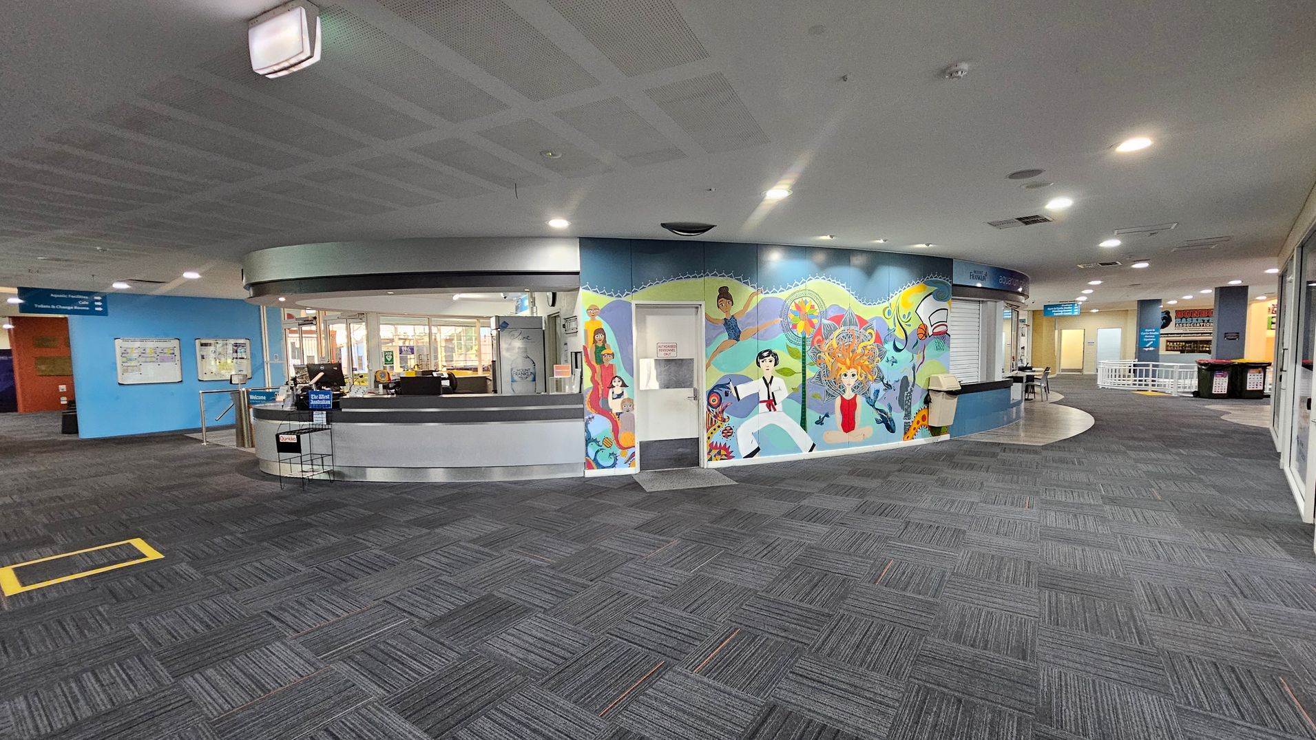 A spacious indoor area with a grey carpeted floor and a ceiling with recessed lighting. On the left, there's a circular reception desk with computers and a "Welcome" sign. Behind it, a blue wall features a colourful mural with various figures and abstract designs. To the right, a corridor leads to other rooms, with signs and a small seating area visible. The overall atmosphere is modern and organised. A spacious indoor area with a grey carpeted floor and a ceiling with recessed lighting. On the left, there's a circular reception desk with computers and a "Welcome" sign. Behind it, a blue wall features a colourful mural with various figures and abstract designs. To the right, a corridor leads to other rooms, with signs and a small seating area visible. The overall atmosphere is modern and organised.