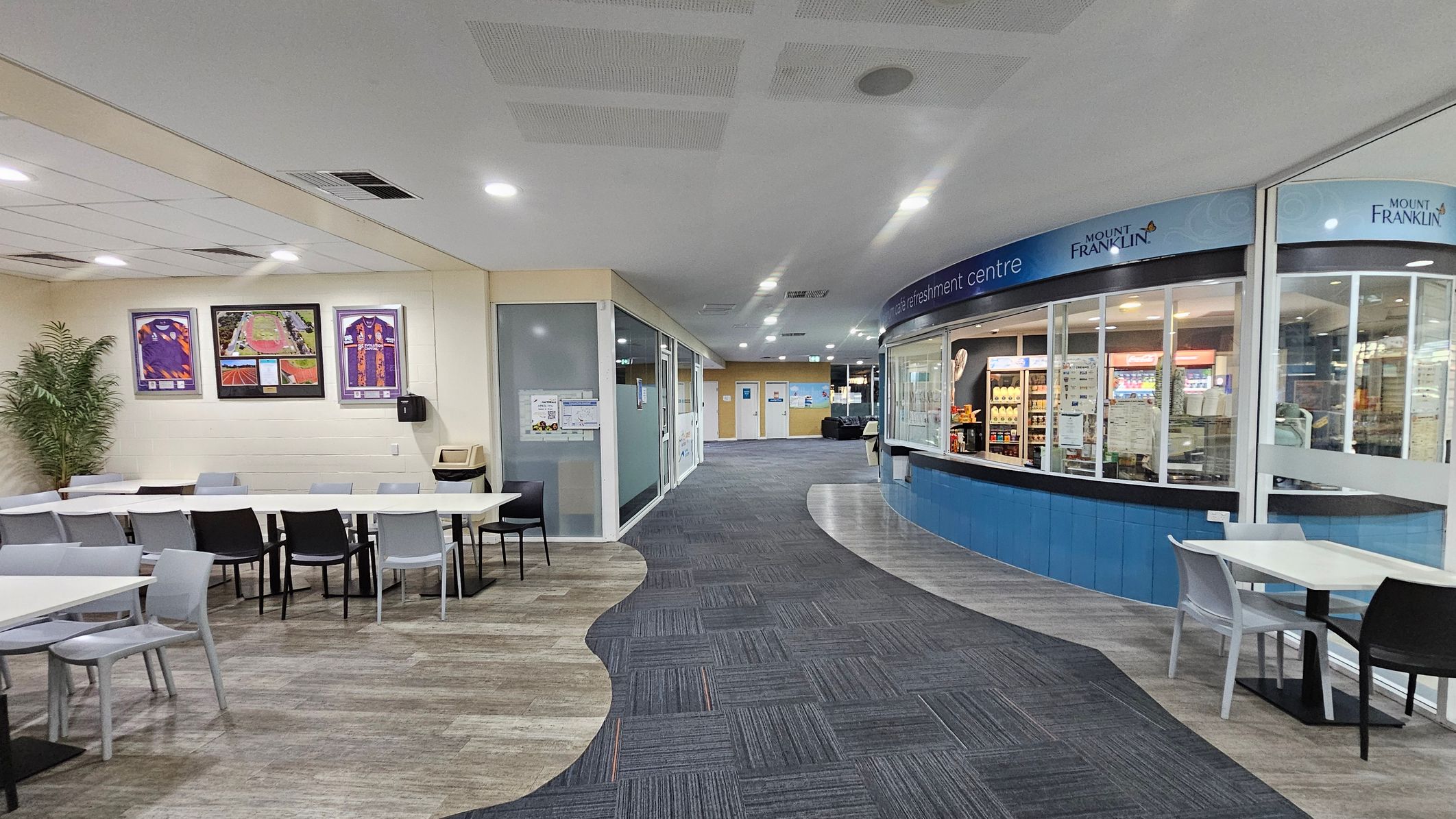 On the left, there are white tables with a mix of black and grey chairs, and framed pictures on the wall above. A potted plant is in the corner. The floor has a mix of green and brown tiles. On the right, there's a refreshment centre with a curved counter and glass display, featuring various drinks and snacks. The floor transitions to grey tiles. The ceiling is white with recessed lighting. In the background, there are doors and a hallway leading further inside. On the left, there are white tables with a mix of black and grey chairs, and framed pictures on the wall above. A potted plant is in the corner. The floor has a mix of green and brown tiles. On the right, there's a refreshment centre with a curved counter and glass display, featuring various drinks and snacks. The floor transitions to grey tiles. The ceiling is white with recessed lighting. In the background, there are doors and a hallway leading further inside.