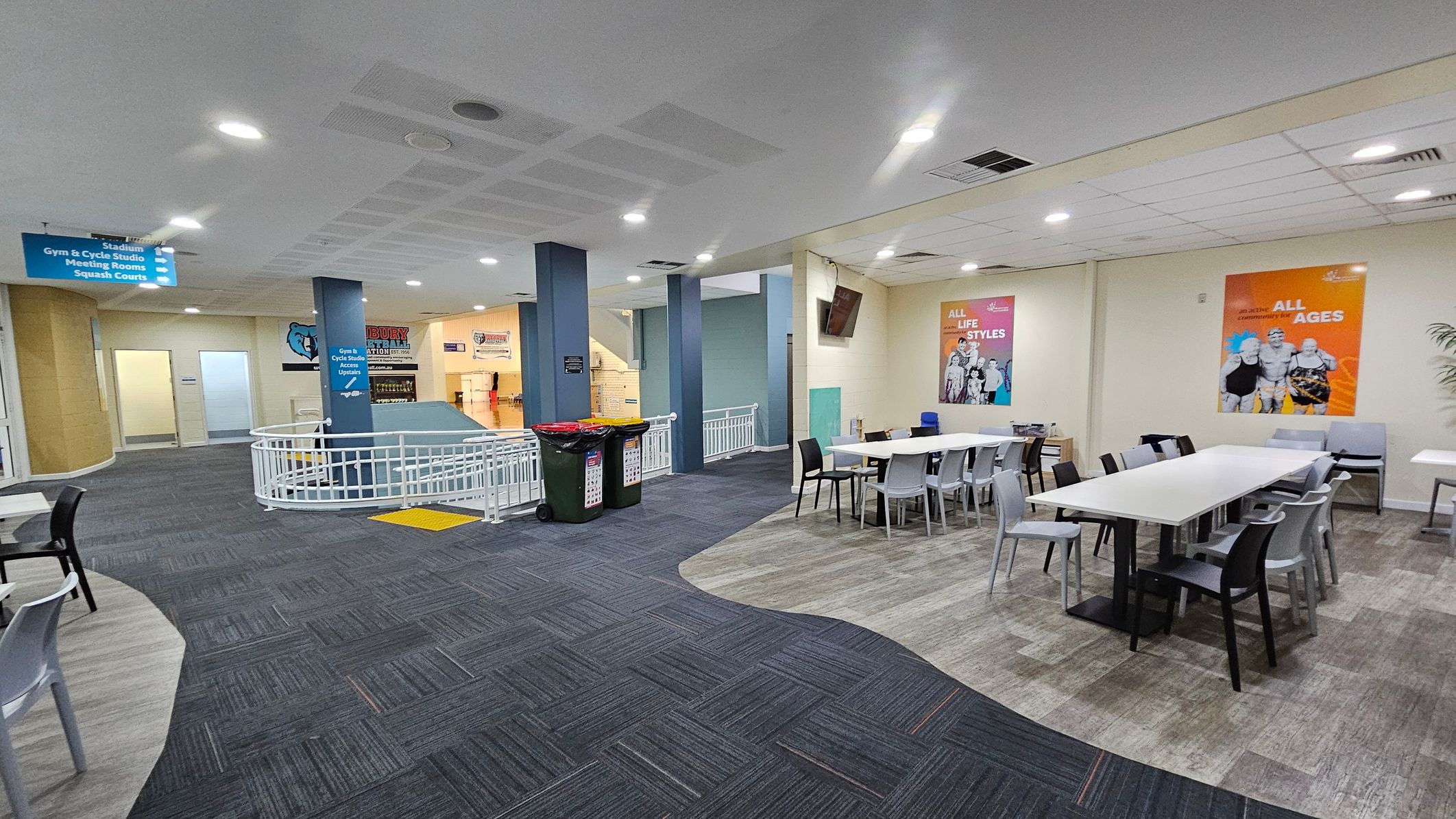 A spacious indoor area with a mix of grey and purple carpeted flooring. On the right, there are white tables surrounded by black and grey chairs, with colourful posters on the wall above. In the centre, a blue pillar with directional signs and a railing around a ramp. On the left, a corridor leads to other rooms, and there are bins near the pillar. The ceiling is fitted with recessed lighting. A spacious indoor area with a mix of grey and purple carpeted flooring. On the right, there are white tables surrounded by black and grey chairs, with colourful posters on the wall above. In the centre, a blue pillar with directional signs and a railing around a ramp. On the left, a corridor leads to other rooms, and there are bins near the pillar. The ceiling is fitted with recessed lighting.