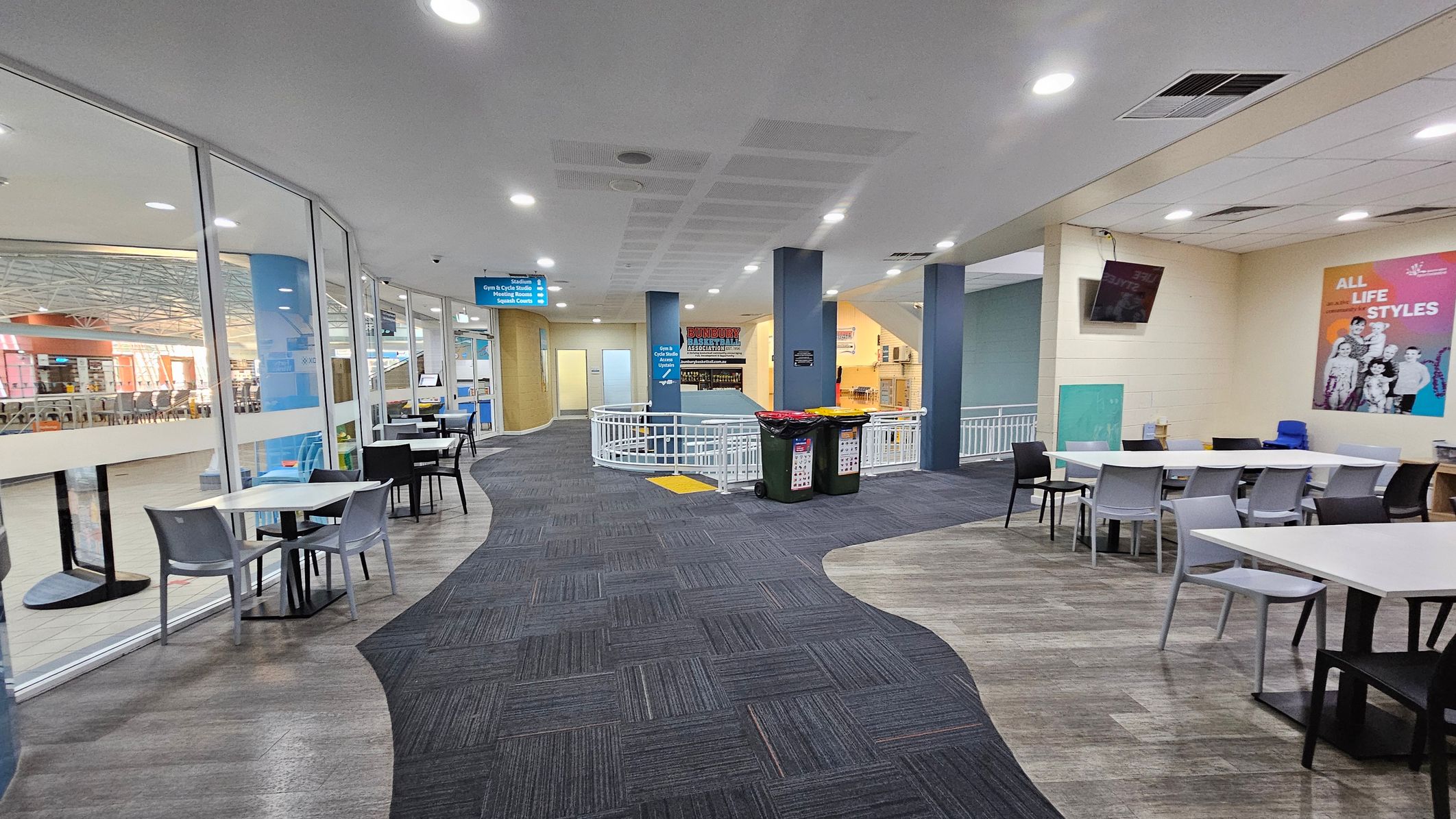 A spacious indoor area with a curved pathway featuring a mix of grey and purple carpet. On the left, there are glass walls with tables and chairs set against them. The right side has more tables and chairs, with a colourful poster on the wall and a mounted TV. In the centre, two bins are placed near a white railing. The ceiling is white with recessed lighting, and there are signs hanging from it. The floor transitions from carpet to a wood-like texture. A spacious indoor area with a curved pathway featuring a mix of grey and purple carpet. On the left, there are glass walls with tables and chairs set against them. The right side has more tables and chairs, with a colourful poster on the wall and a mounted TV. In the centre, two bins are placed near a white railing. The ceiling is white with recessed lighting, and there are signs hanging from it. The floor transitions from carpet to a wood-like texture.