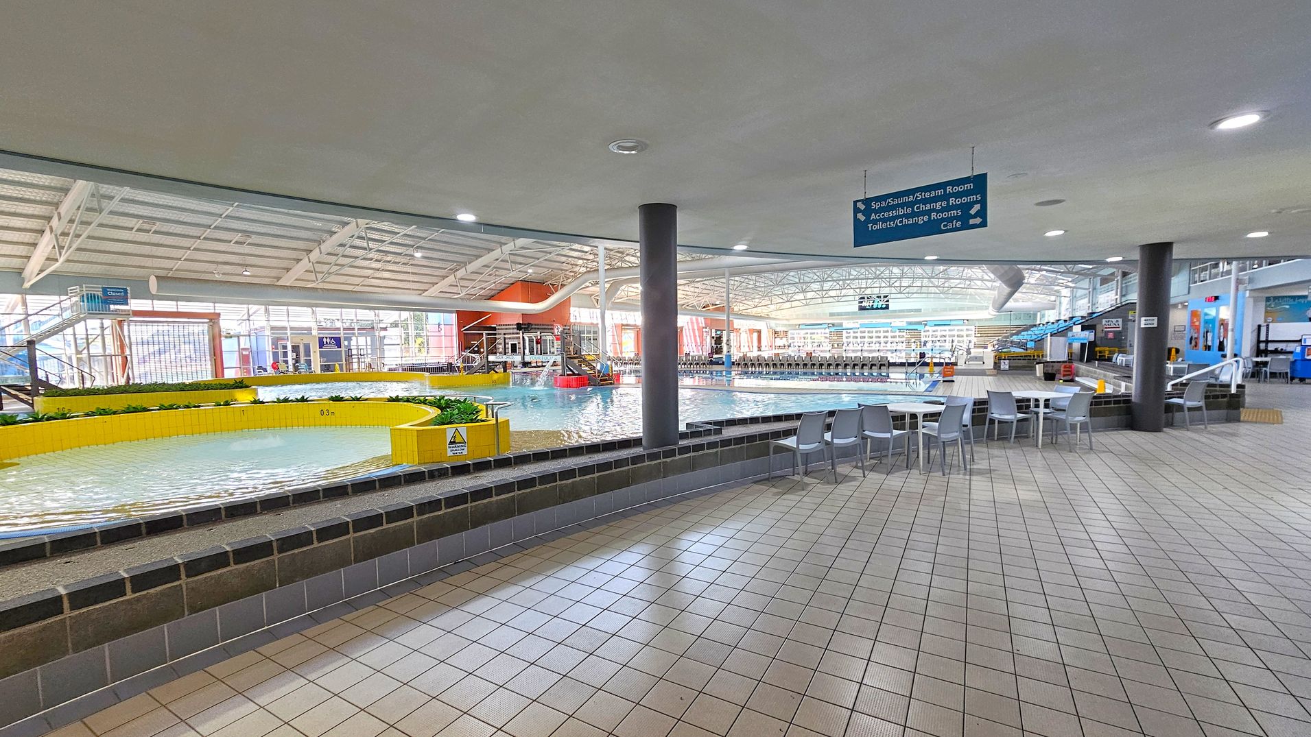 An indoor swimming pool area with a tiled floor. On the left, there's a shallow circular pool with yellow tiled edges and some plants. In the centre, a larger pool with clear water is visible, surrounded by a spacious area with tables and chairs. The ceiling is high with metal beams and lights. On the right, there are more seating areas and signs indicating directions to facilities like a spa, sauna, and café. The background shows large windows and additional pool features. An indoor swimming pool area with a tiled floor. On the left, there's a shallow circular pool with yellow tiled edges and some plants. In the centre, a larger pool with clear water is visible, surrounded by a spacious area with tables and chairs. The ceiling is high with metal beams and lights. On the right, there are more seating areas and signs indicating directions to facilities like a spa, sauna, and café. The background shows large windows and additional pool features.