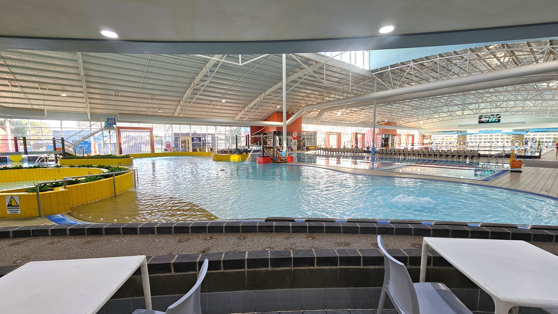 Indoor swimming pool area with a shallow pool in the foreground, surrounded by a low black-tiled wall. White tables and chairs are positioned near the pool. The pool has a yellow-tiled circular feature with plants. In the background, there are more pools, a slide, and a row of chairs. The ceiling is high with metal beams and skylights, allowing natural light to illuminate the space. Indoor swimming pool area with a shallow pool in the foreground, surrounded by a low black-tiled wall. White tables and chairs are positioned near the pool. The pool has a yellow-tiled circular feature with plants. In the background, there are more pools, a slide, and a row of chairs. The ceiling is high with metal beams and skylights, allowing natural light to illuminate the space.