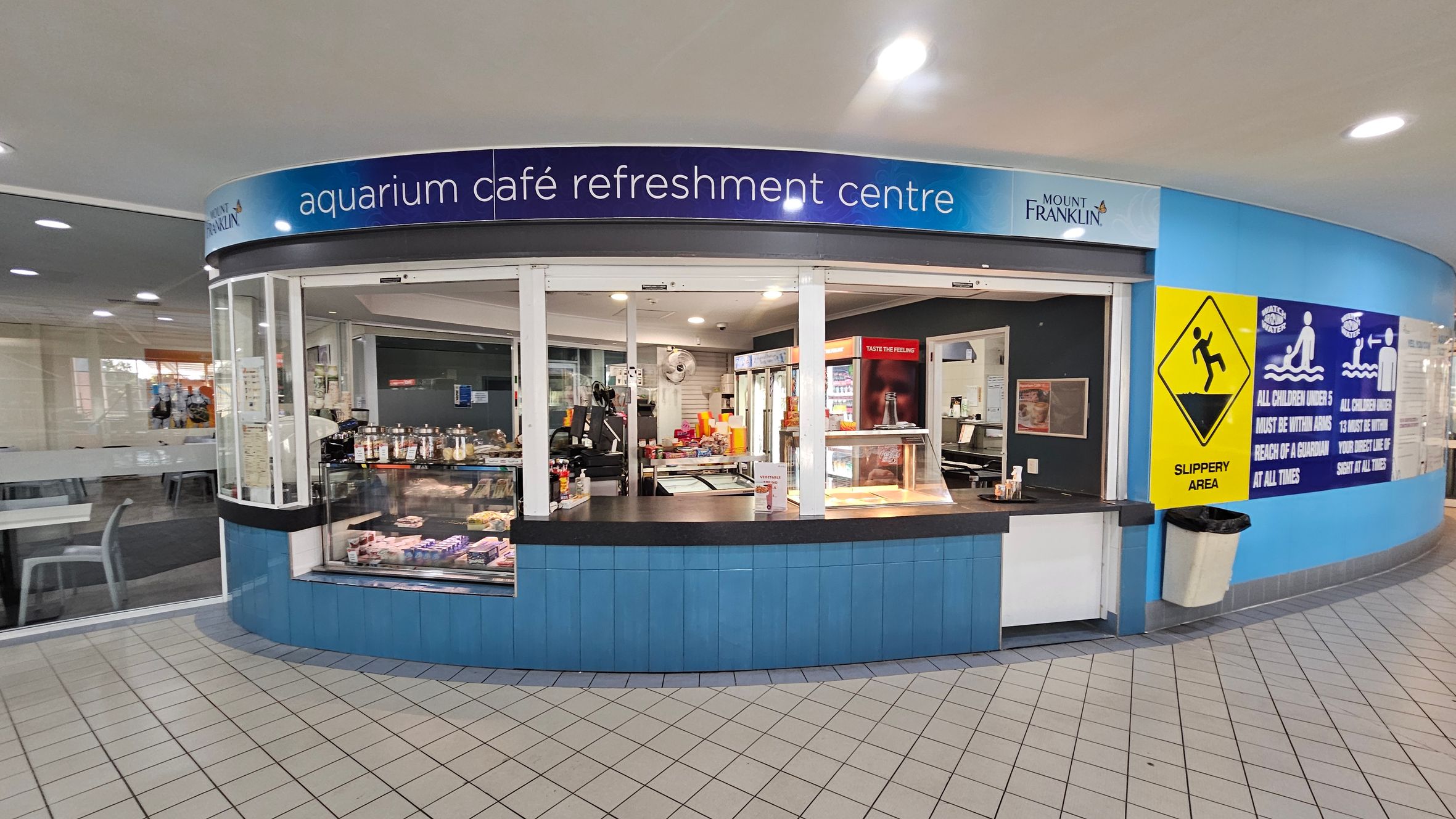 Aquarium café refreshment centre with a curved counter displaying snacks and drinks. The counter is blue with a black top. Behind the counter, there are shelves with various food items and a drinks fridge. On the right, a yellow "Slippery Area" sign and blue safety notices are visible. The floor is tiled in a light colour, and the ceiling has recessed lighting. To the left, there are tables and chairs in a seating area. Aquarium café refreshment centre with a curved counter displaying snacks and drinks. The counter is blue with a black top. Behind the counter, there are shelves with various food items and a drinks fridge. On the right, a yellow "Slippery Area" sign and blue safety notices are visible. The floor is tiled in a light colour, and the ceiling has recessed lighting. To the left, there are tables and chairs in a seating area.