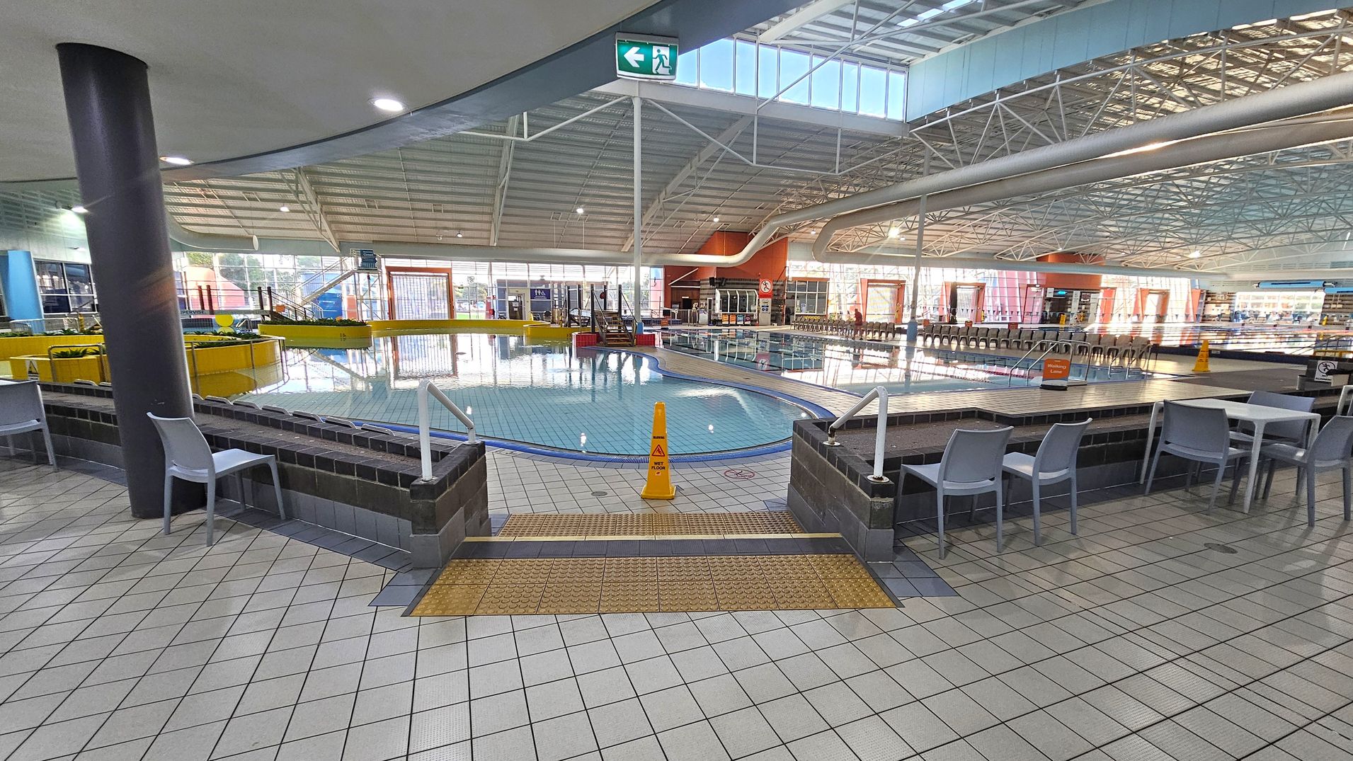 Indoor swimming pool area with a tiled floor. In the foreground, there are tables and chairs on either side. A small pool with a yellow safety cone is in the centre, surrounded by a low brick wall. The background features a larger pool with a slide and a row of chairs along the far wall. The ceiling is high with metal beams and large windows allowing natural light. An exit sign is visible above. Indoor swimming pool area with a tiled floor. In the foreground, there are tables and chairs on either side. A small pool with a yellow safety cone is in the centre, surrounded by a low brick wall. The background features a larger pool with a slide and a row of chairs along the far wall. The ceiling is high with metal beams and large windows allowing natural light. An exit sign is visible above.