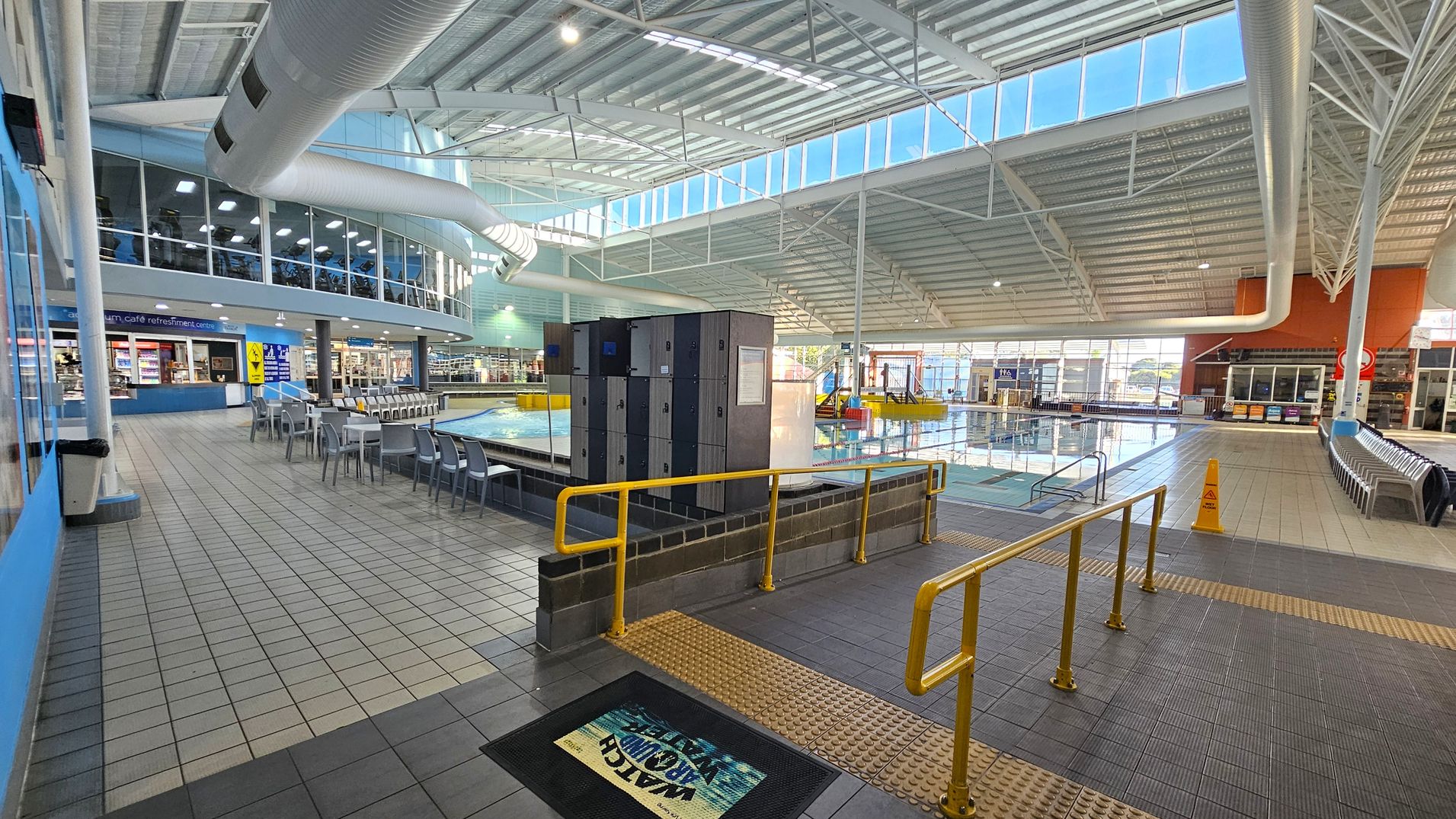 An indoor aquatic centre with a large pool on the right, surrounded by tiled flooring. In the foreground, there are yellow railings and a mat with a warning sign. Lockers are positioned near the pool. On the left, a café area with tables and chairs is visible, and above it, a mezzanine with exercise equipment. The ceiling is high with exposed beams and large windows allowing natural light to enter. An indoor aquatic centre with a large pool on the right, surrounded by tiled flooring. In the foreground, there are yellow railings and a mat with a warning sign. Lockers are positioned near the pool. On the left, a café area with tables and chairs is visible, and above it, a mezzanine with exercise equipment. The ceiling is high with exposed beams and large windows allowing natural light to enter.