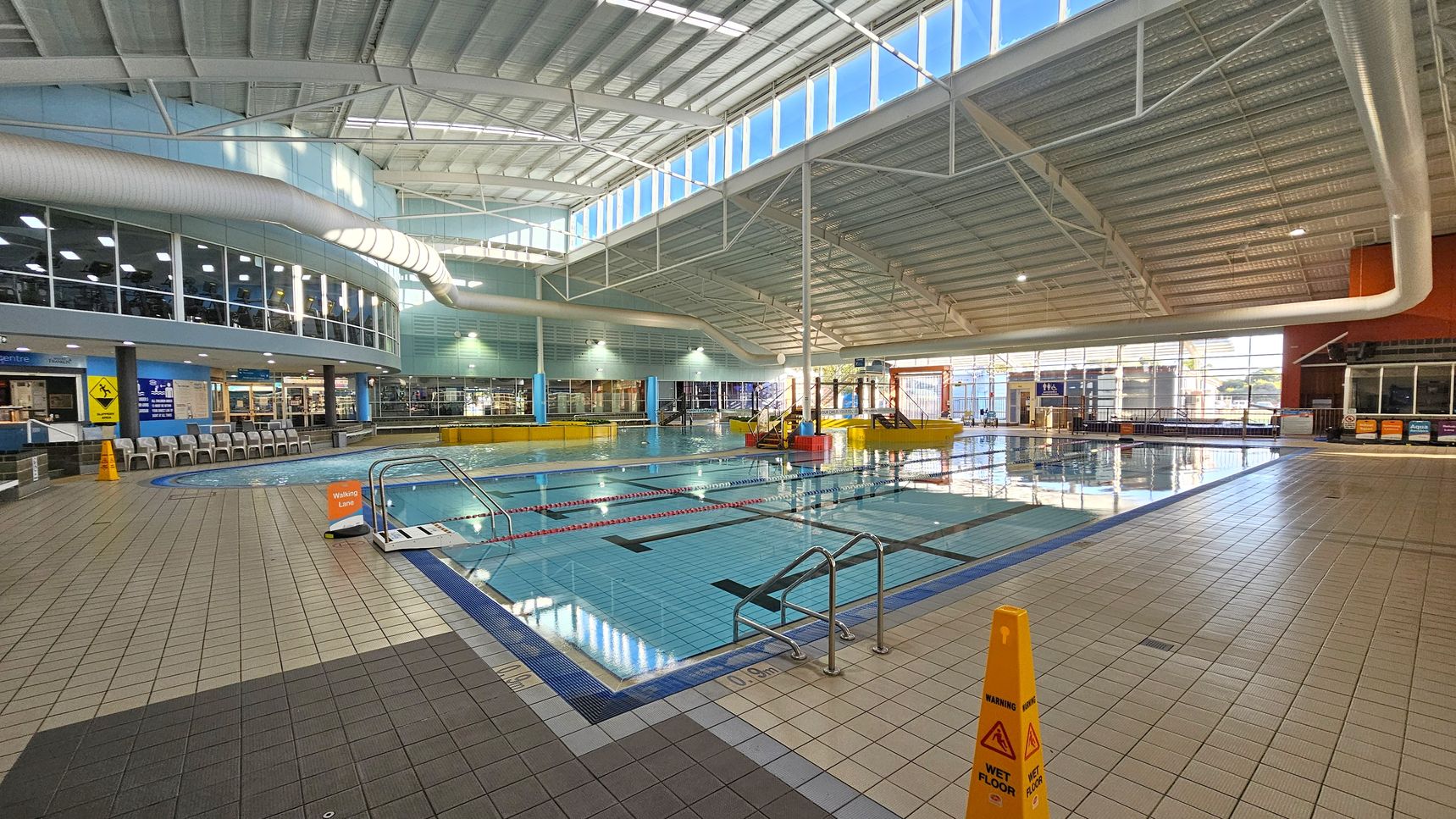 An indoor swimming pool area with a tiled floor and a high, curved ceiling featuring large windows allowing natural light. On the left, there are rows of chairs and a glass-walled gym area above. In the centre, the pool has marked lanes and a small set of steps leading into the water. On the right, there are more windows and a play area with water features. Yellow caution signs are placed around the pool, indicating a wet floor. An indoor swimming pool area with a tiled floor and a high, curved ceiling featuring large windows allowing natural light. On the left, there are rows of chairs and a glass-walled gym area above. In the centre, the pool has marked lanes and a small set of steps leading into the water. On the right, there are more windows and a play area with water features. Yellow caution signs are placed around the pool, indicating a wet floor.