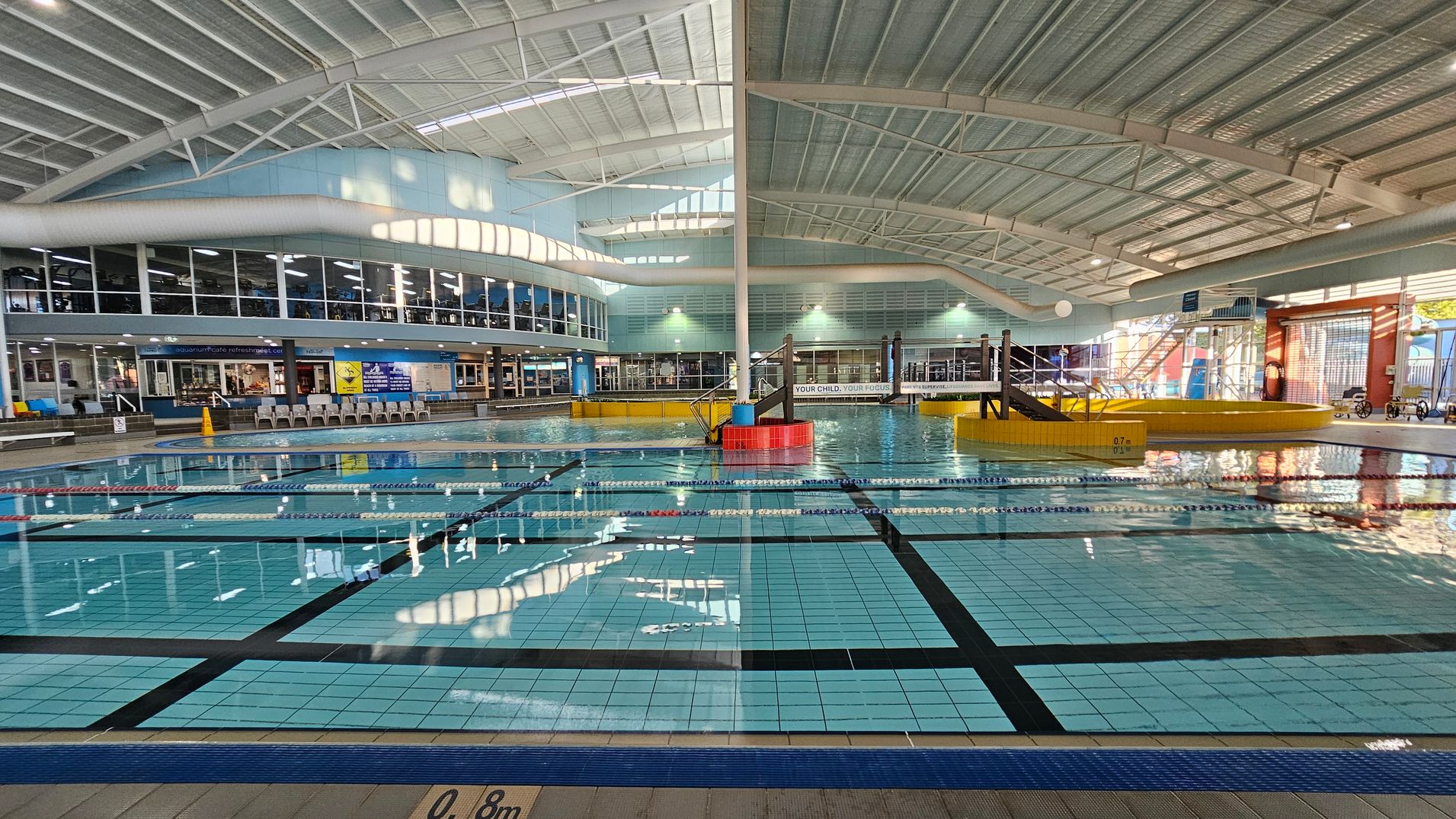 Indoor swimming pool with clear blue water and lane markings. On the left, a row of lounge chairs is visible near large windows. In the centre, a small slide and play area are surrounded by a yellow barrier. The ceiling is high with metal beams and skylights. The right side has more windows and a staircase leading to an upper level. The pool depth is marked at 0.8 metres. Indoor swimming pool with clear blue water and lane markings. On the left, a row of lounge chairs is visible near large windows. In the centre, a small slide and play area are surrounded by a yellow barrier. The ceiling is high with metal beams and skylights. The right side has more windows and a staircase leading to an upper level. The pool depth is marked at 0.8 metres.