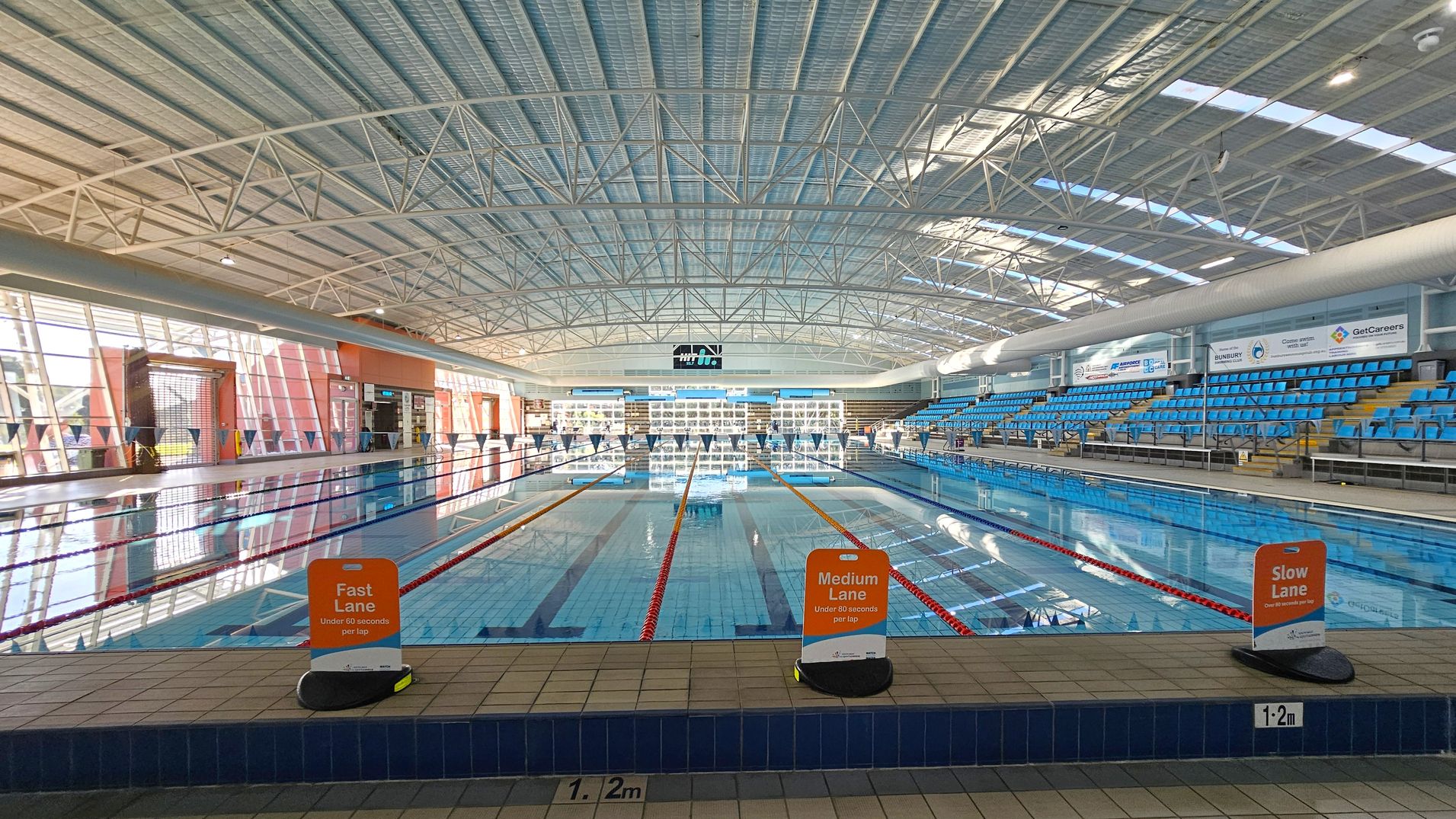 An indoor swimming pool with three lanes marked by signs: "Fast Lane," "Medium Lane," and "Slow Lane." The pool is surrounded by blue seating on the right side. The ceiling is high with a metal framework and skylights. Large windows on the left allow natural light to enter. The pool deck is tiled, and the water is clear, reflecting the structure above. Signs and advertisements are visible on the walls. An indoor swimming pool with three lanes marked by signs: "Fast Lane," "Medium Lane," and "Slow Lane." The pool is surrounded by blue seating on the right side. The ceiling is high with a metal framework and skylights. Large windows on the left allow natural light to enter. The pool deck is tiled, and the water is clear, reflecting the structure above. Signs and advertisements are visible on the walls.