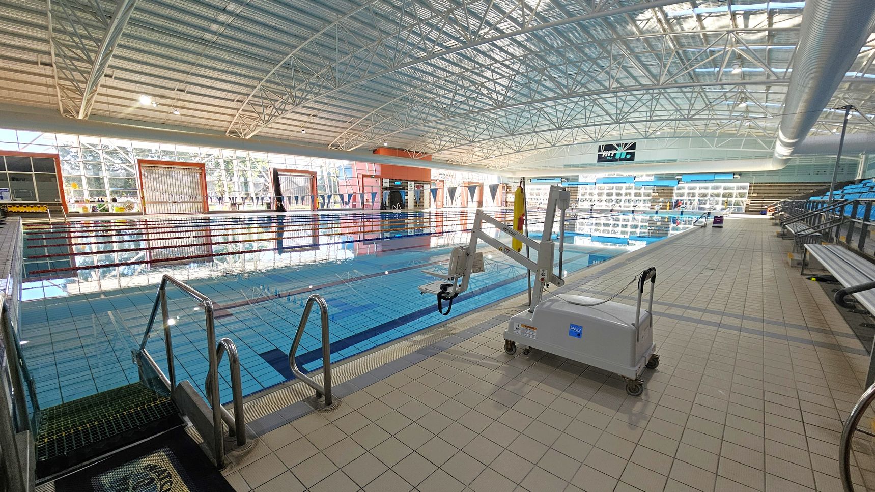 An indoor swimming pool with clear blue water and lane dividers. On the right, there's a pool lift device on the tiled deck. The left side features metal handrails leading into the pool. In the background, large windows allow natural light to illuminate the space, and there are rows of blue seating for spectators. The ceiling is high with a metal framework and a large ventilation duct running along the right side. An indoor swimming pool with clear blue water and lane dividers. On the right, there's a pool lift device on the tiled deck. The left side features metal handrails leading into the pool. In the background, large windows allow natural light to illuminate the space, and there are rows of blue seating for spectators. The ceiling is high with a metal framework and a large ventilation duct running along the right side.
