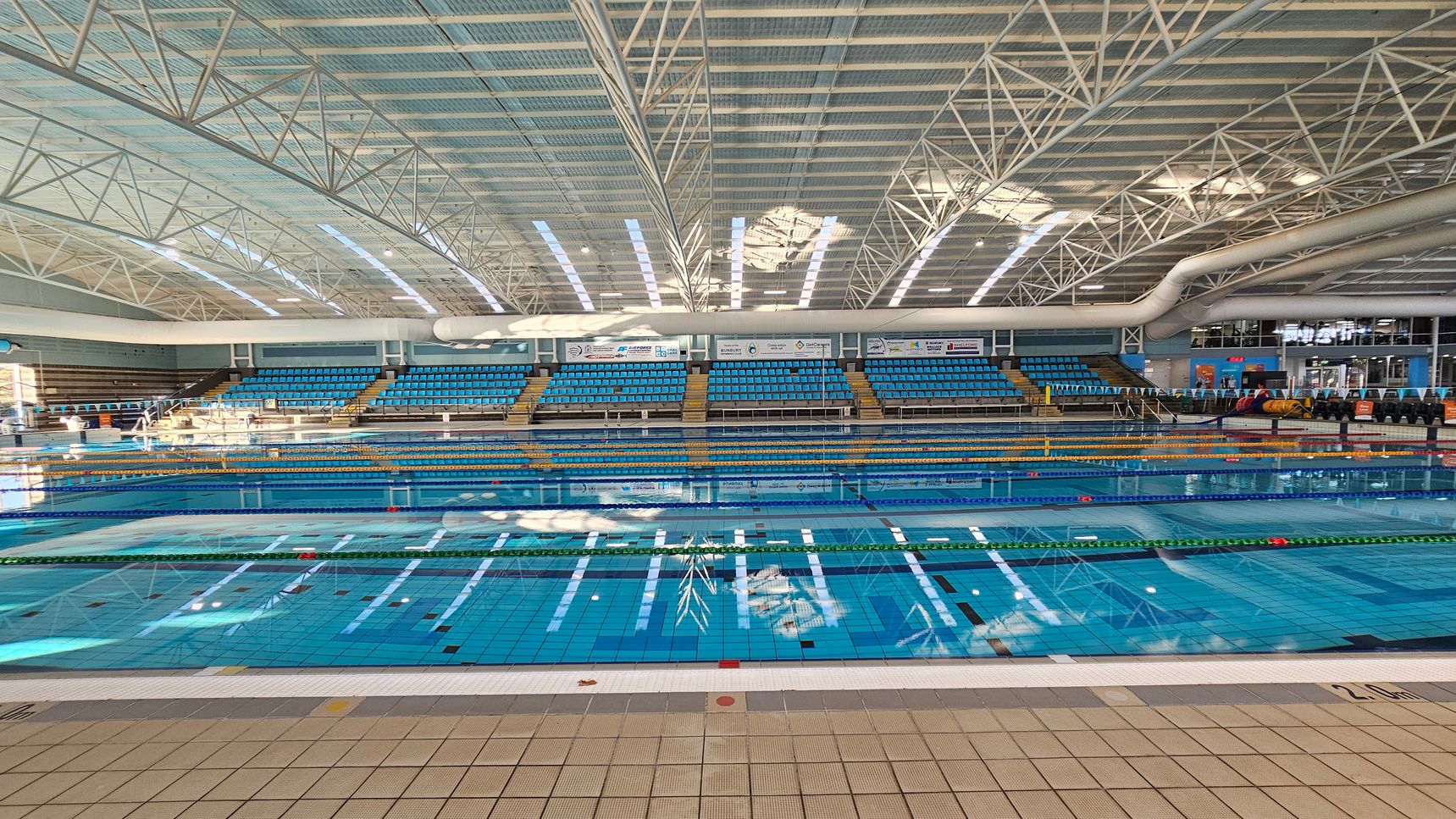An indoor swimming pool with clear blue water, divided into lanes by floating lane markers. The background features tiered seating with blue chairs, and a large scoreboard or display area above. The ceiling is high with a grid of metal beams and skylights allowing natural light to filter through. The floor around the pool is tiled in a light colour. An indoor swimming pool with clear blue water, divided into lanes by floating lane markers. The background features tiered seating with blue chairs, and a large scoreboard or display area above. The ceiling is high with a grid of metal beams and skylights allowing natural light to filter through. The floor around the pool is tiled in a light colour.