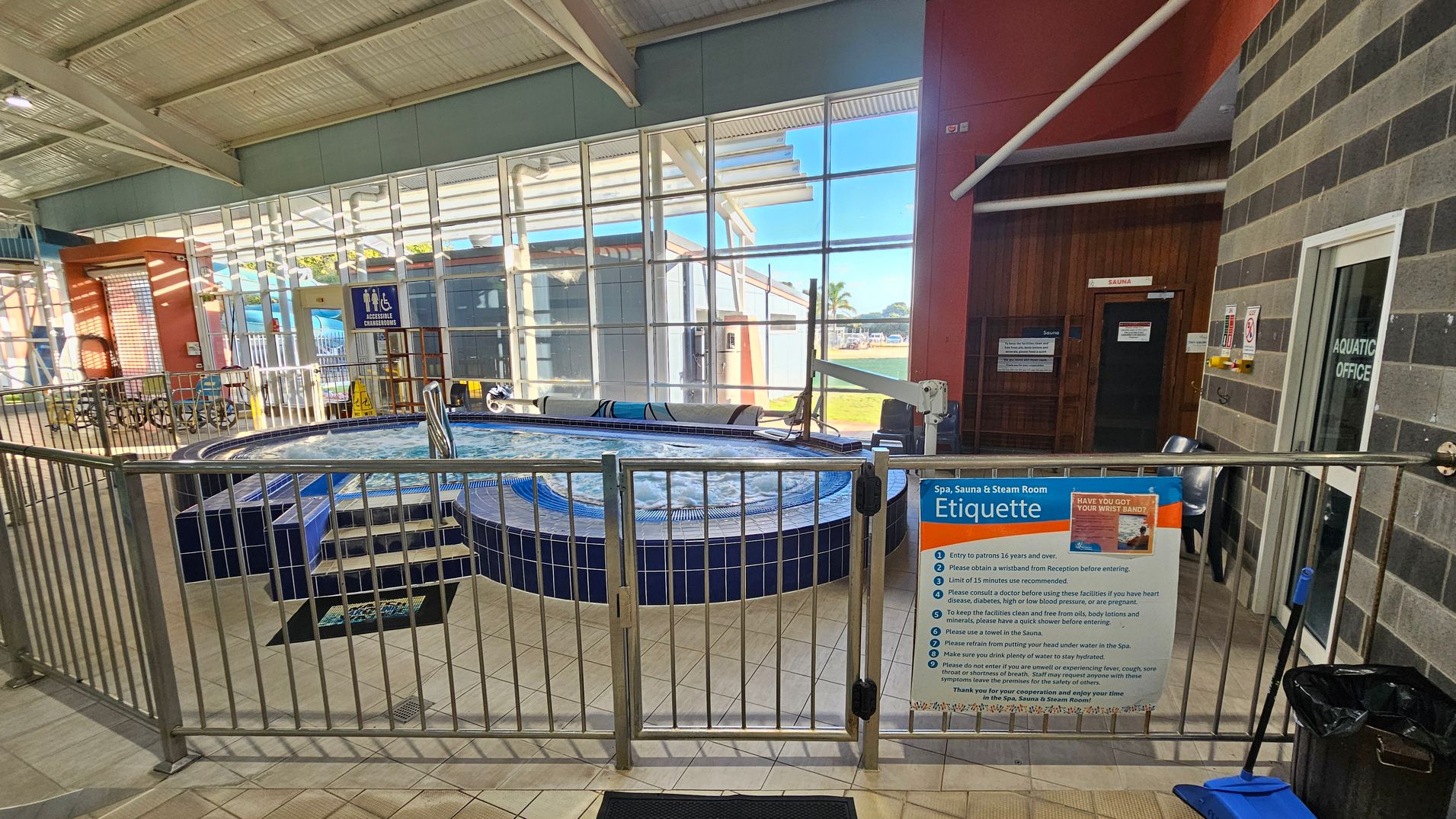A spa area with a circular hot tub surrounded by a metal fence. On the right, a sign displays spa etiquette rules. Behind the fence, a door marked "Sauna" is visible. The left side shows large windows with a view of the outside, allowing natural light to illuminate the space. The floor is tiled, and a blue dustpan and brush are placed near the fence. The ceiling is made of metal panels with exposed beams. A spa area with a circular hot tub surrounded by a metal fence. On the right, a sign displays spa etiquette rules. Behind the fence, a door marked "Sauna" is visible. The left side shows large windows with a view of the outside, allowing natural light to illuminate the space. The floor is tiled, and a blue dustpan and brush are placed near the fence. The ceiling is made of metal panels with exposed beams.